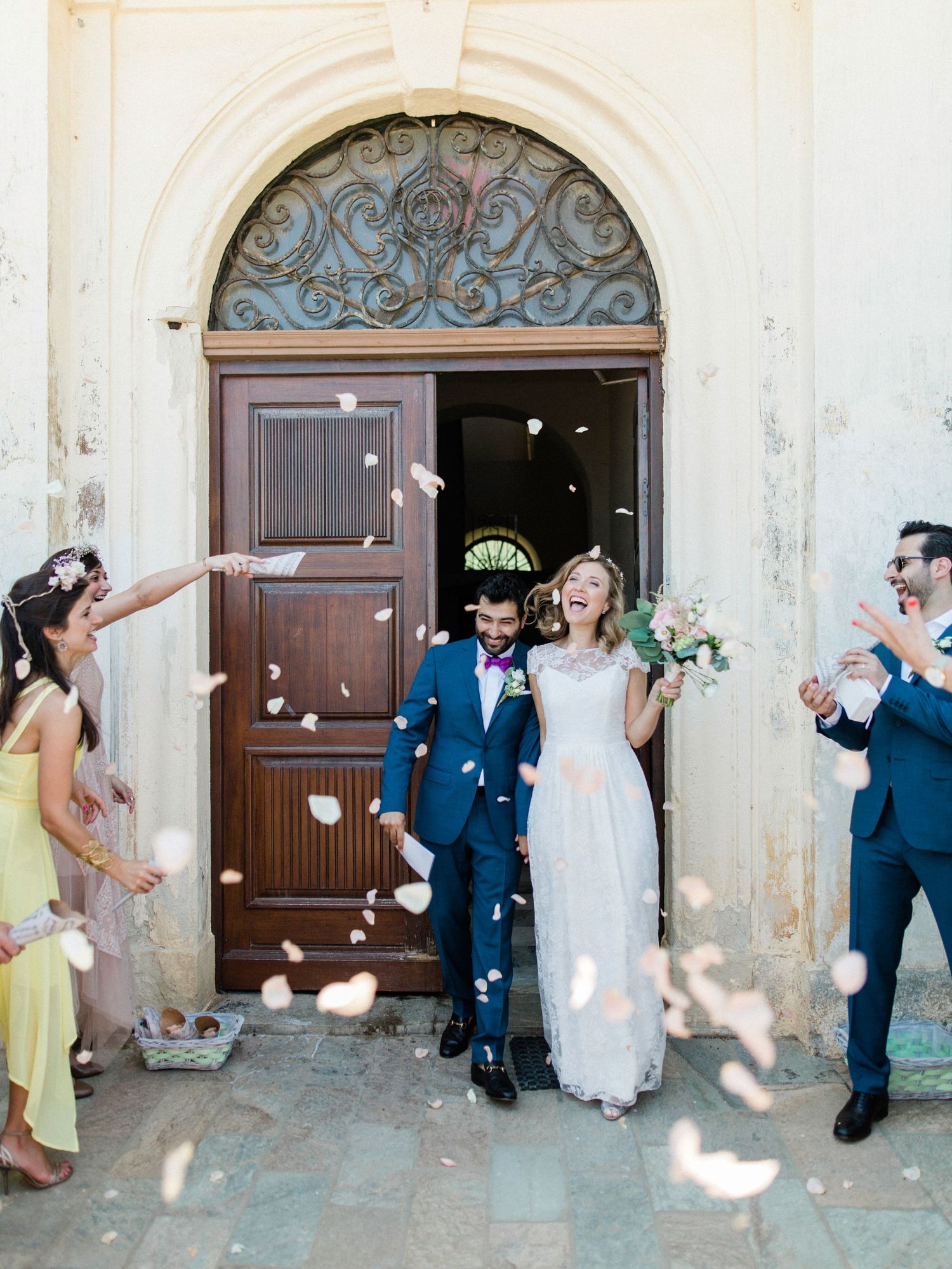 Joyful newlyweds exit church doors as guests toss flower petal confetti, bride in white gown, groom in navy suit.