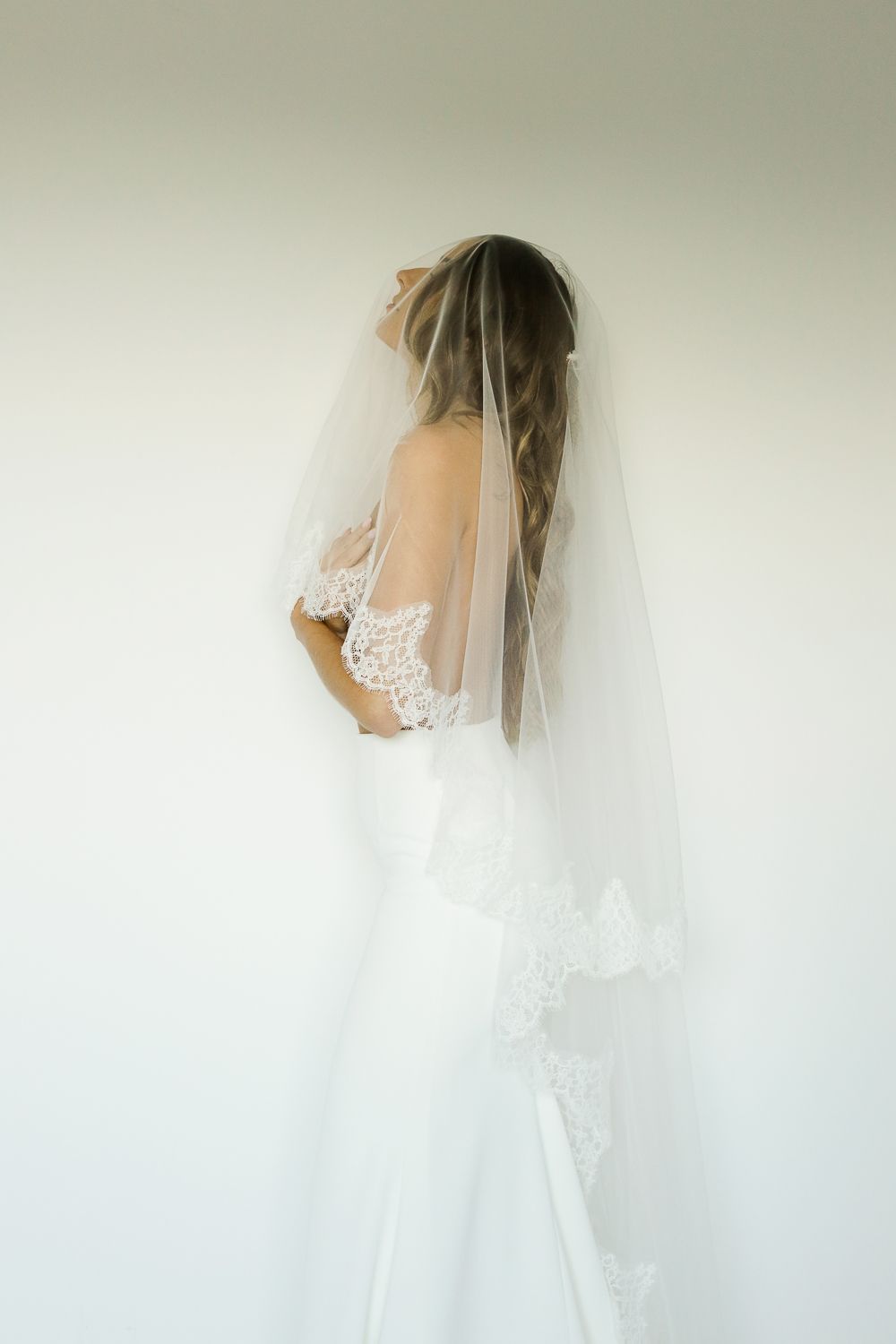 Bride in white lace wedding dress and sheer veil with lace trim, viewed from behind against a light wall.
