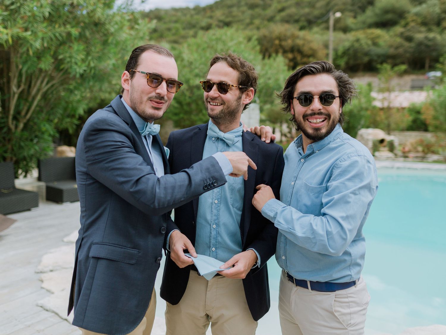 Trois hommes portant des lunettes de soleil sourient et posent ensemble près d'une piscine lors d'un événement en plein air.