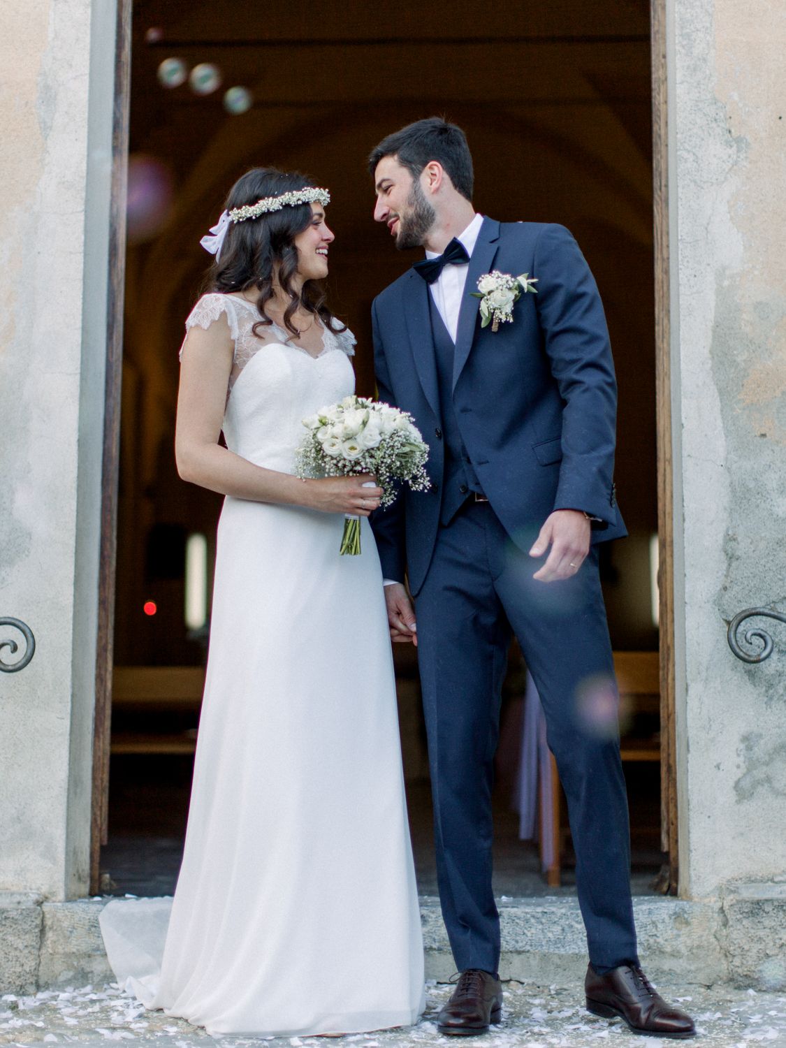 Bride and groom share a romantic moment outside a church, she holds a white bouquet wearing a floral crown.