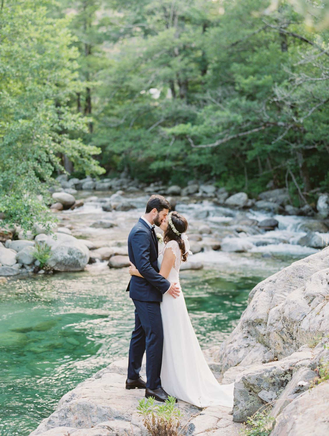 Bride and groom share a romantic kiss on rocky riverbank surrounded by lush green trees and turquoise water.