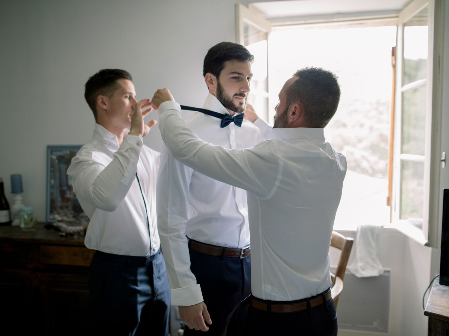 Two groomsmen help a groom adjust his navy bow tie while getting ready for a wedding ceremony indoors.