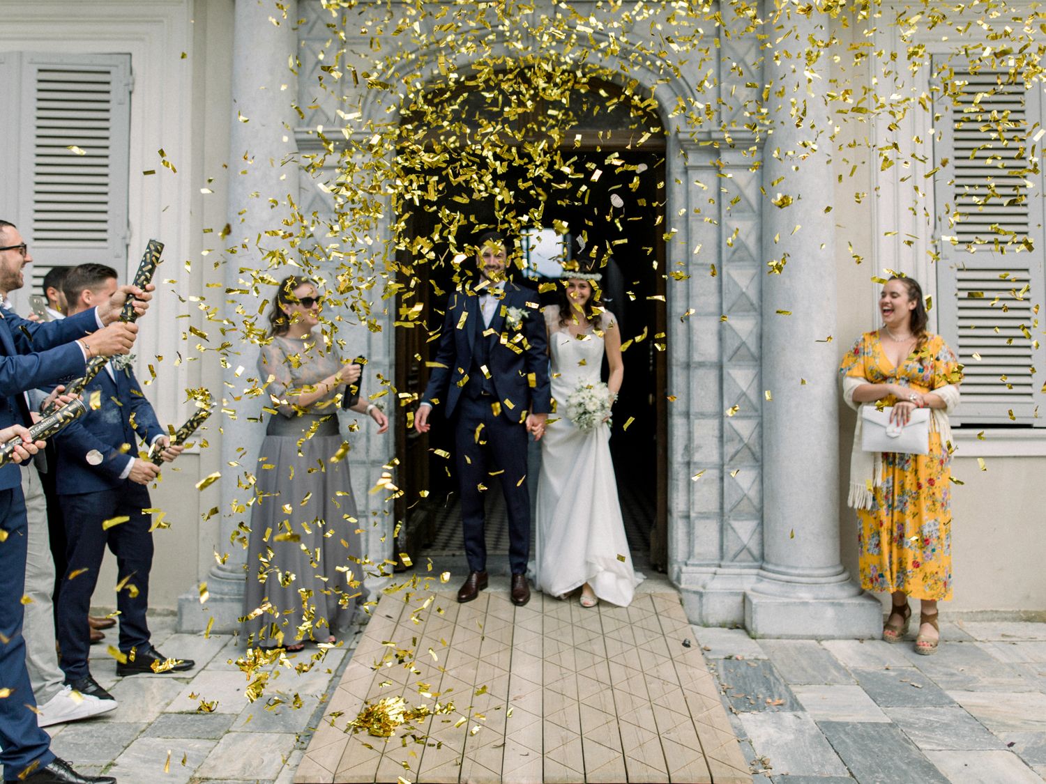 Bride and groom exit church doors as guests celebrate with golden confetti cannons at outdoor wedding ceremony.