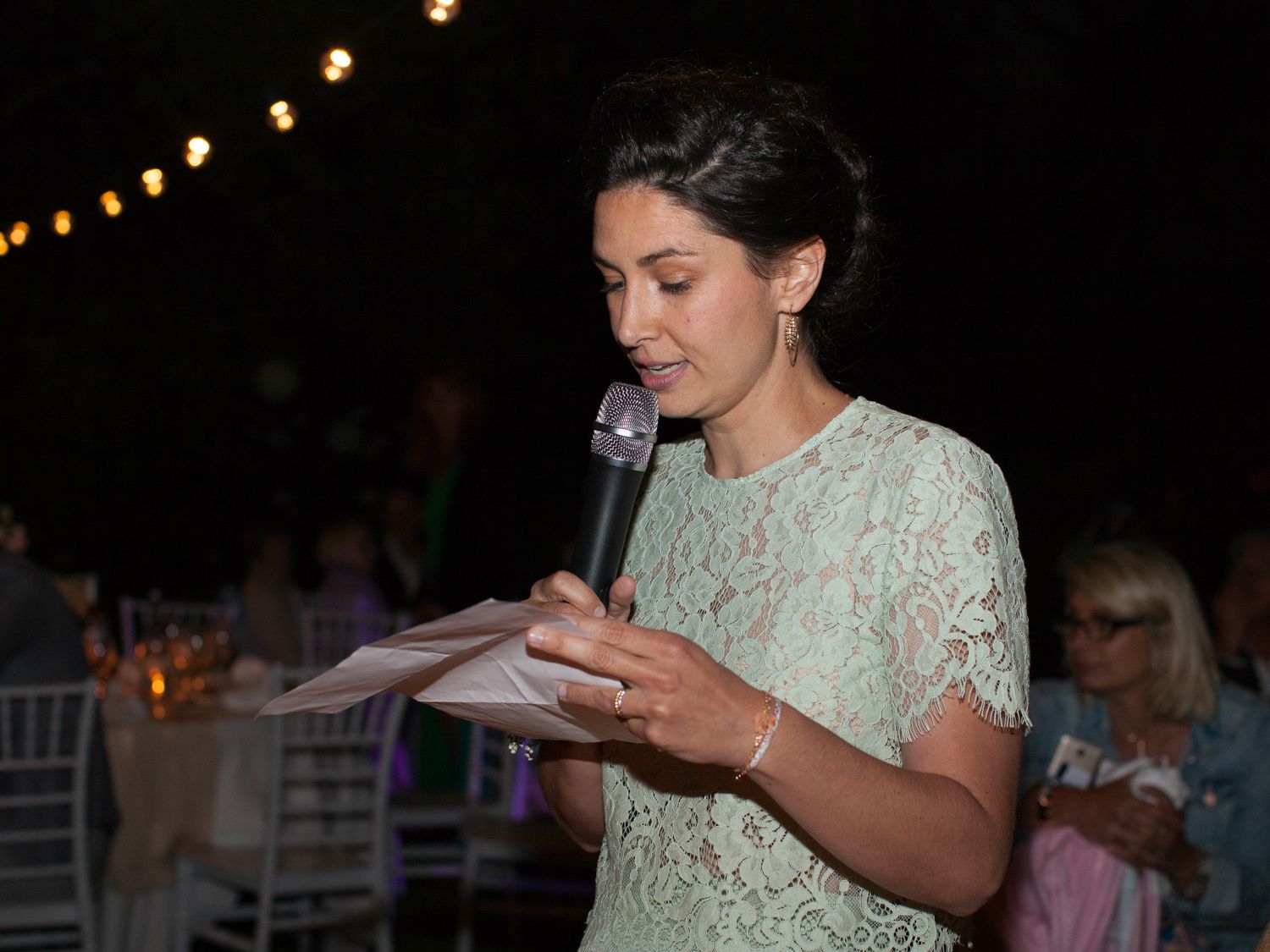 Une femme en robe de dentelle lit un discours au microphone lors d'un événement en plein air en soirée.