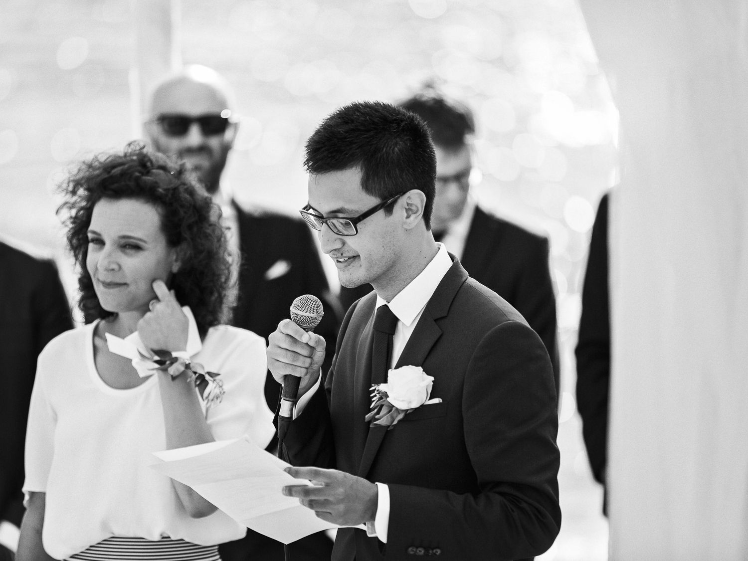 Le marié en costume lit ses vœux lors de la cérémonie de mariage tandis qu'une demoiselle d'honneur se tient à ses côtés, micro en main.