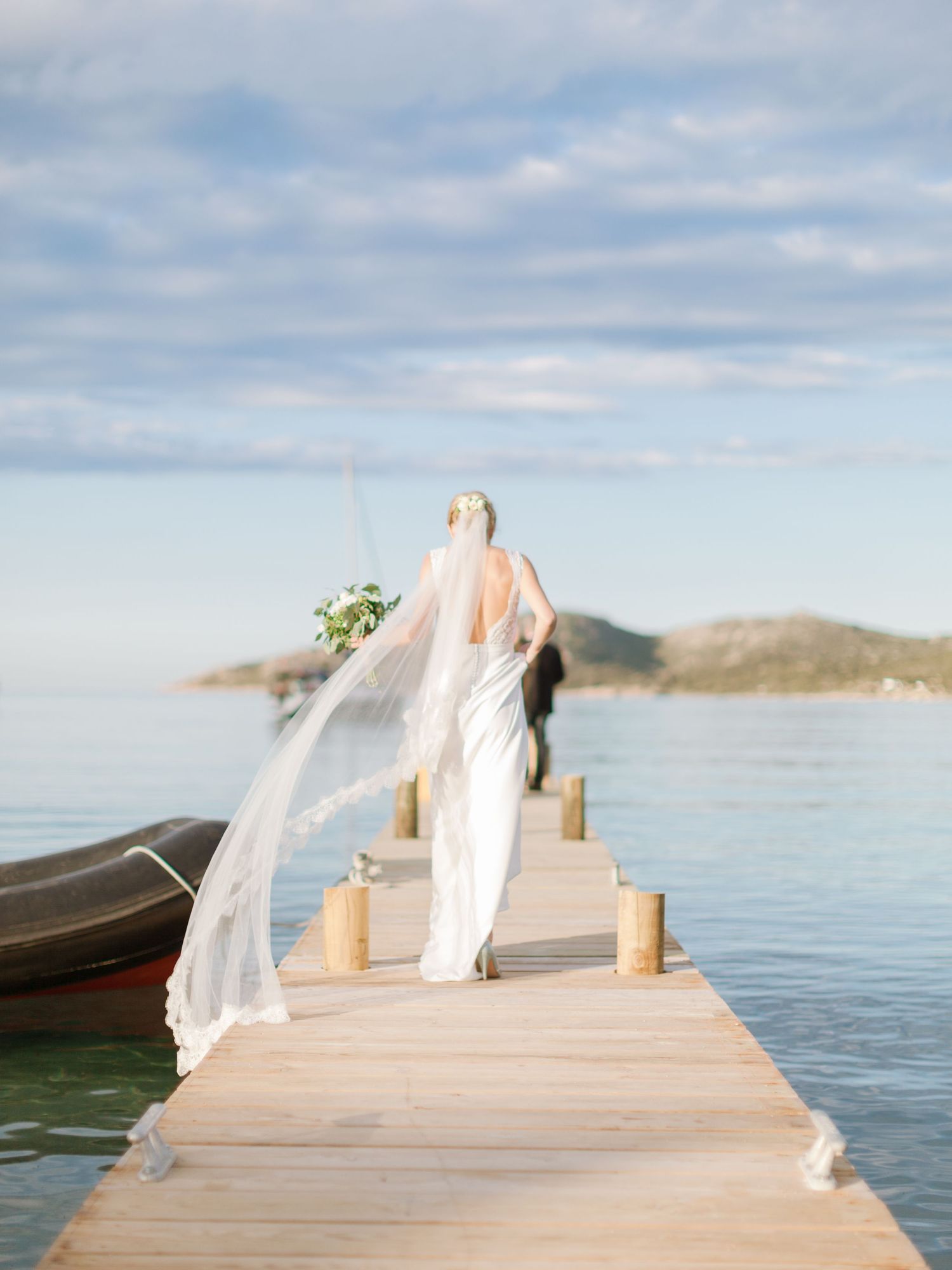 La mariée, vêtue d'une robe blanche et d'un voile fluide, marche sur un quai en bois au-dessus d'une eau turquoise à Bonifacio en corse