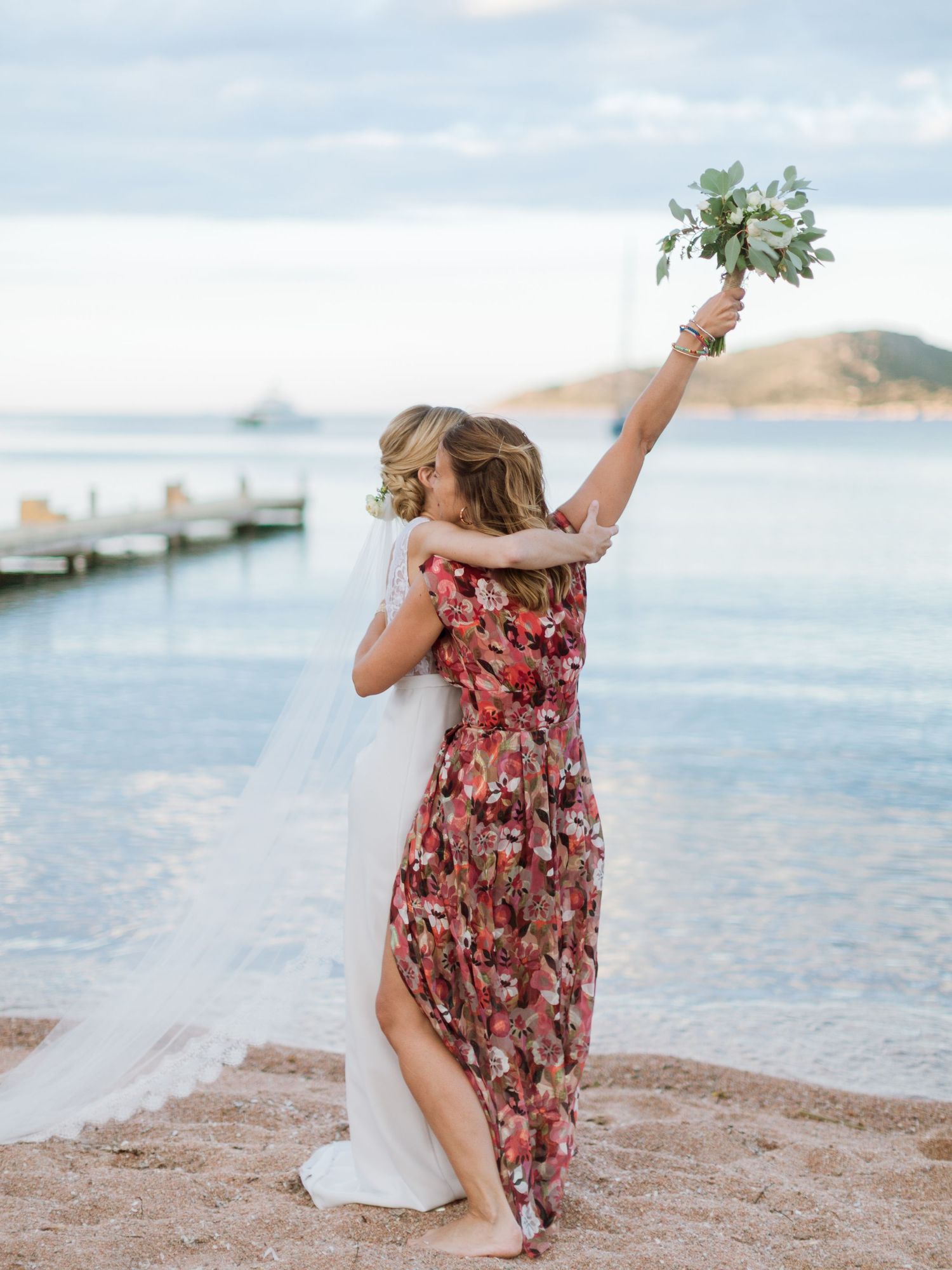 La mariée et son amie s'enlacent sur la plage, la femme en robe à fleurs lève triomphalement son bouquet de mariage