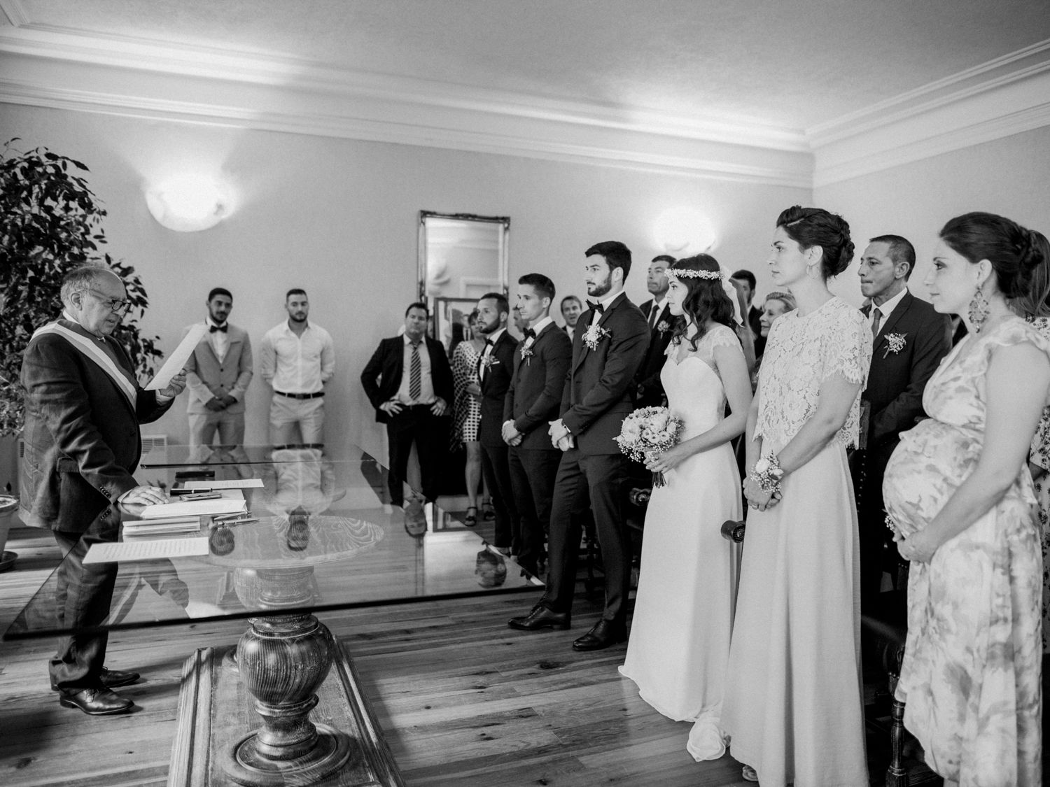 Black and white photo of a wedding ceremony indoors, with bride, groom, and guests gathered before an officiant.