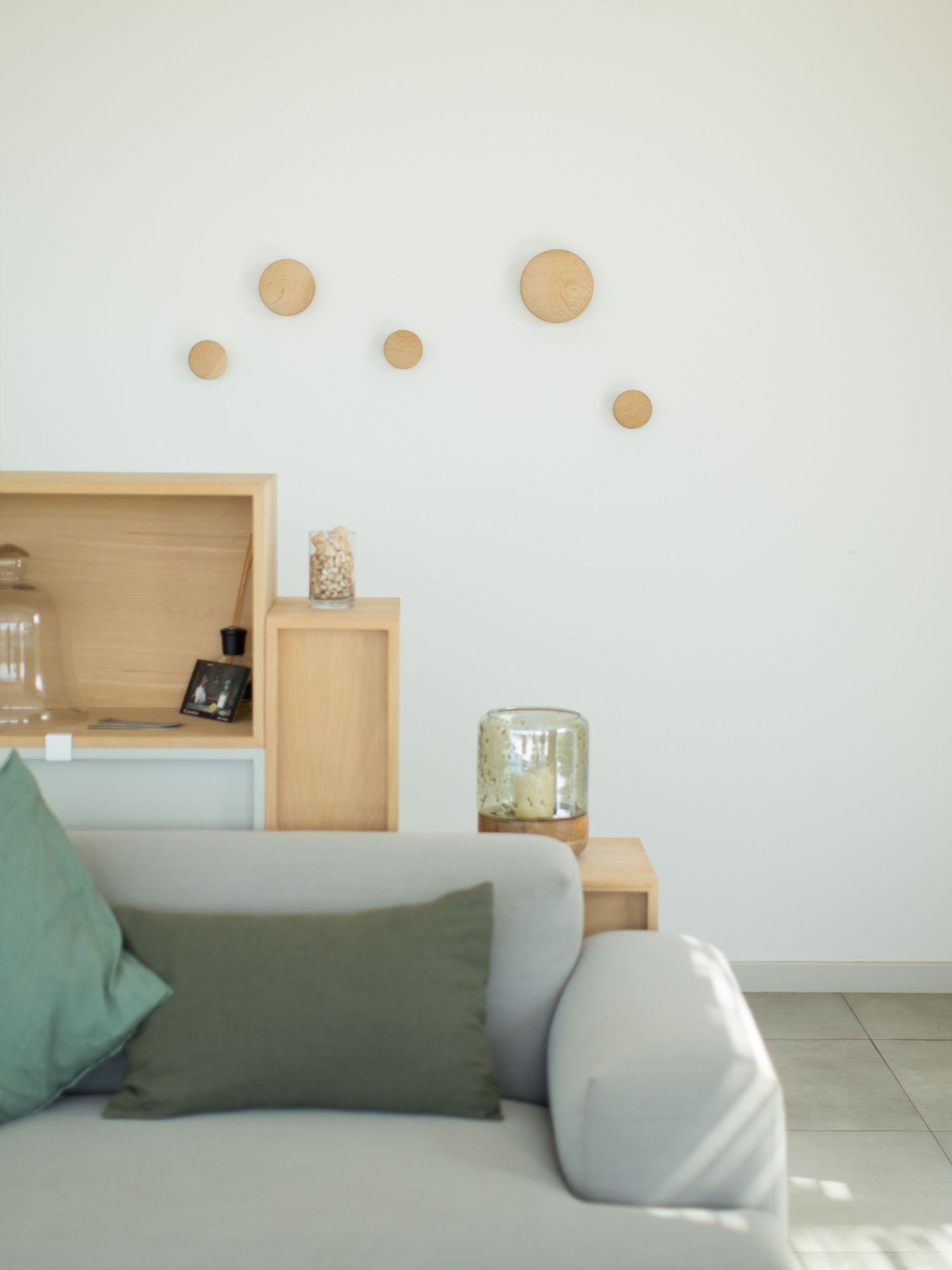 Coin salon minimaliste avec canapé gris clair, coussins verts, étagère en bois et décoration murale circulaire.