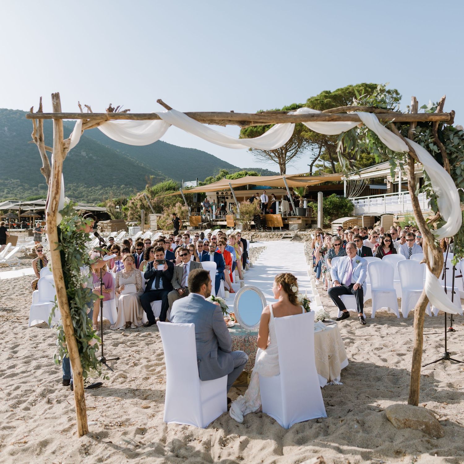 Cérémonie de mariage en plein air sur la plage, avec le couple assis sous une arche en bois rustique ornée de tissu blanc et de feuillage.