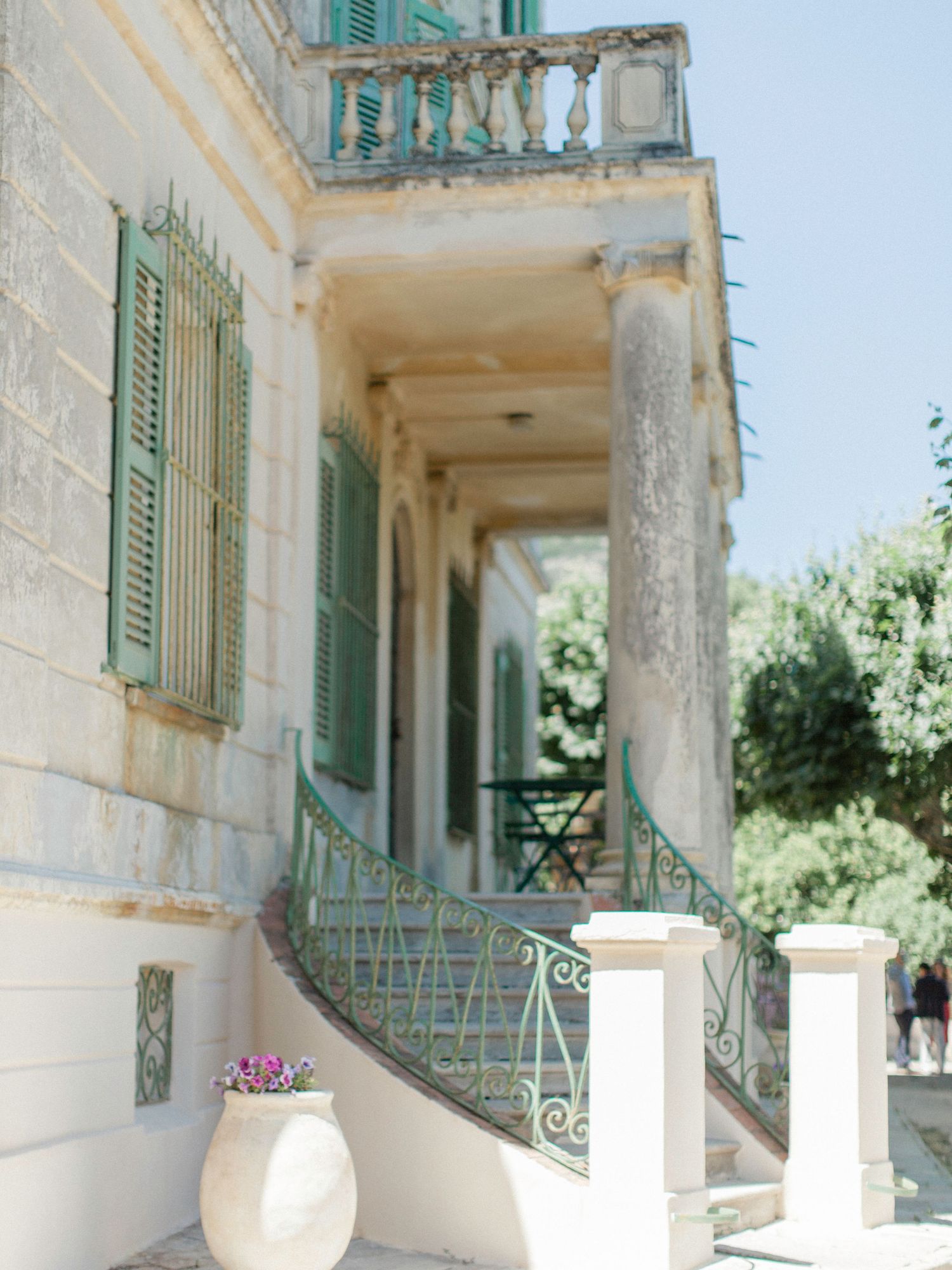 Elegant Mediterranean villa exterior with ornate green iron staircase, shuttered windows, and stone balustrade in warm sunlight.