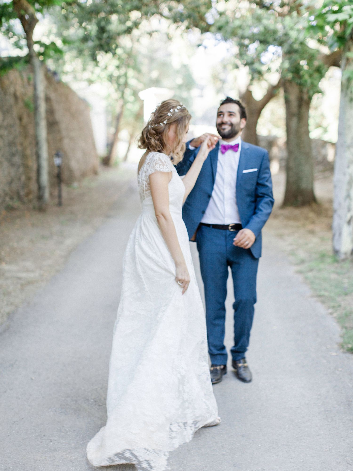 Bride and groom walking on a tree-lined path, she touches his purple bow tie on their wedding day.
