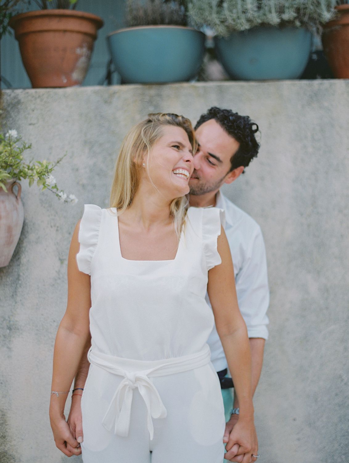 Un couple souriant, vêtu de blanc, partage un tendre moment en plein air ; l'homme embrasse la joue de la femme près de pots en terre cuite