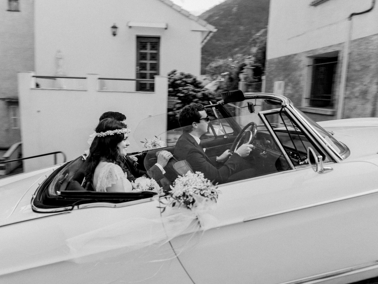 Photo en noir et blanc d'un couple de mariés à bord d'une voiture décapotable blanche classique décorée de fleurs à Corte