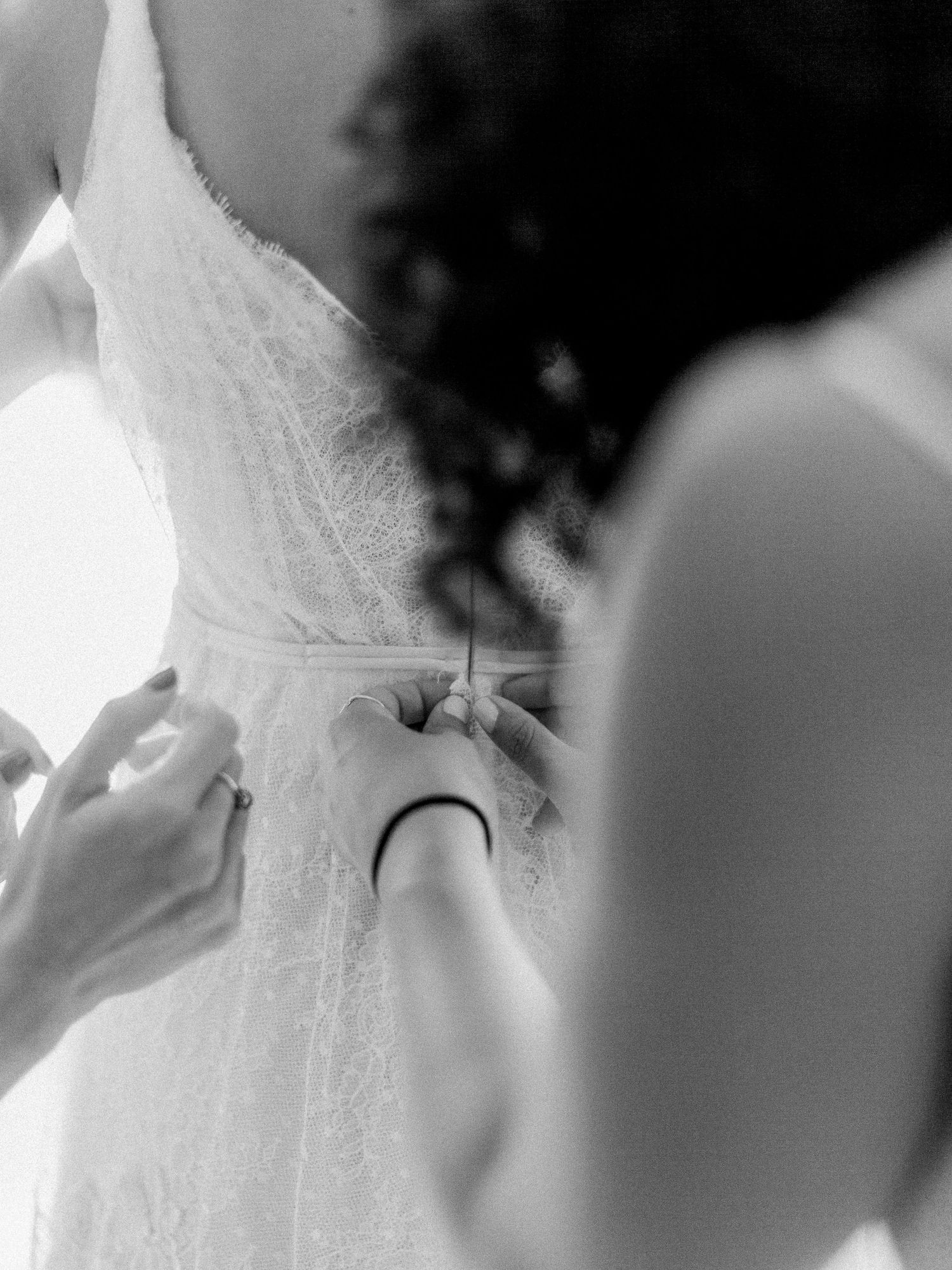 Photo en noir et blanc de demoiselles d'honneur aidant la mariée à attacher sa robe de mariée en dentelle dans le dos.