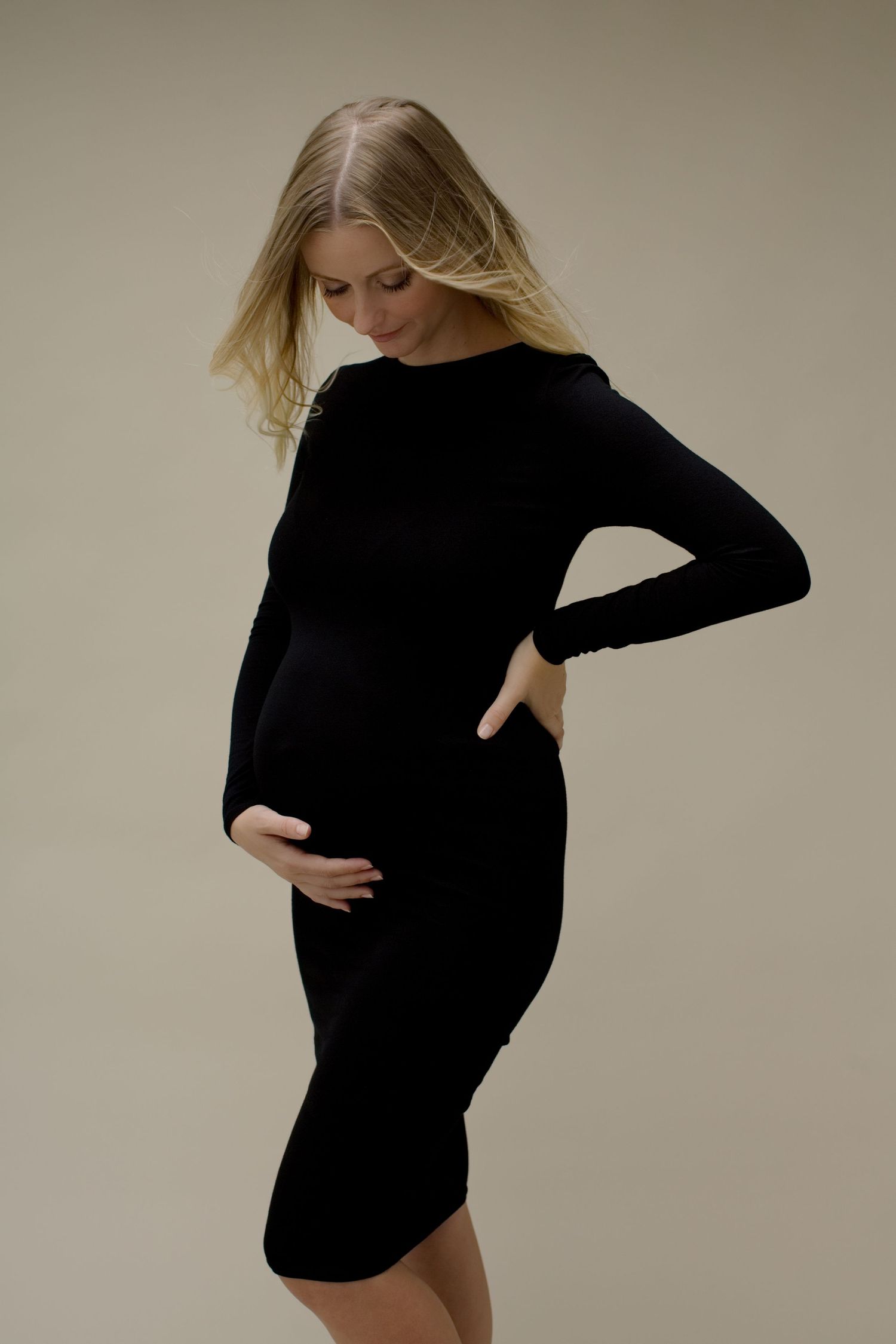 Femme enceinte en robe noire moulante, regardant vers le bas et tenant son ventre rond dans un studio.