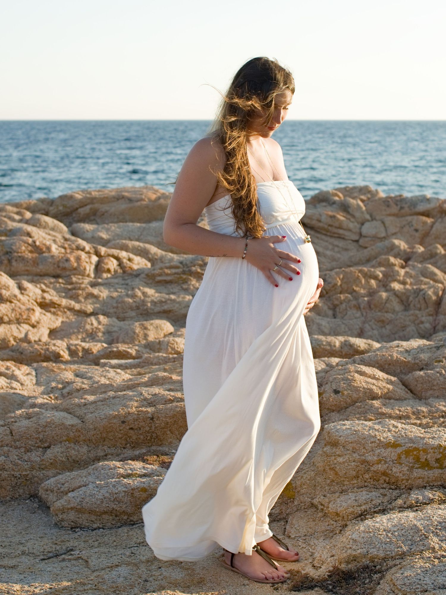 Une femme enceinte, vêtue d'une robe blanche fluide, se tient debout sur des falaises rocheuses en bord de mer, berçant son ventre arrondi.