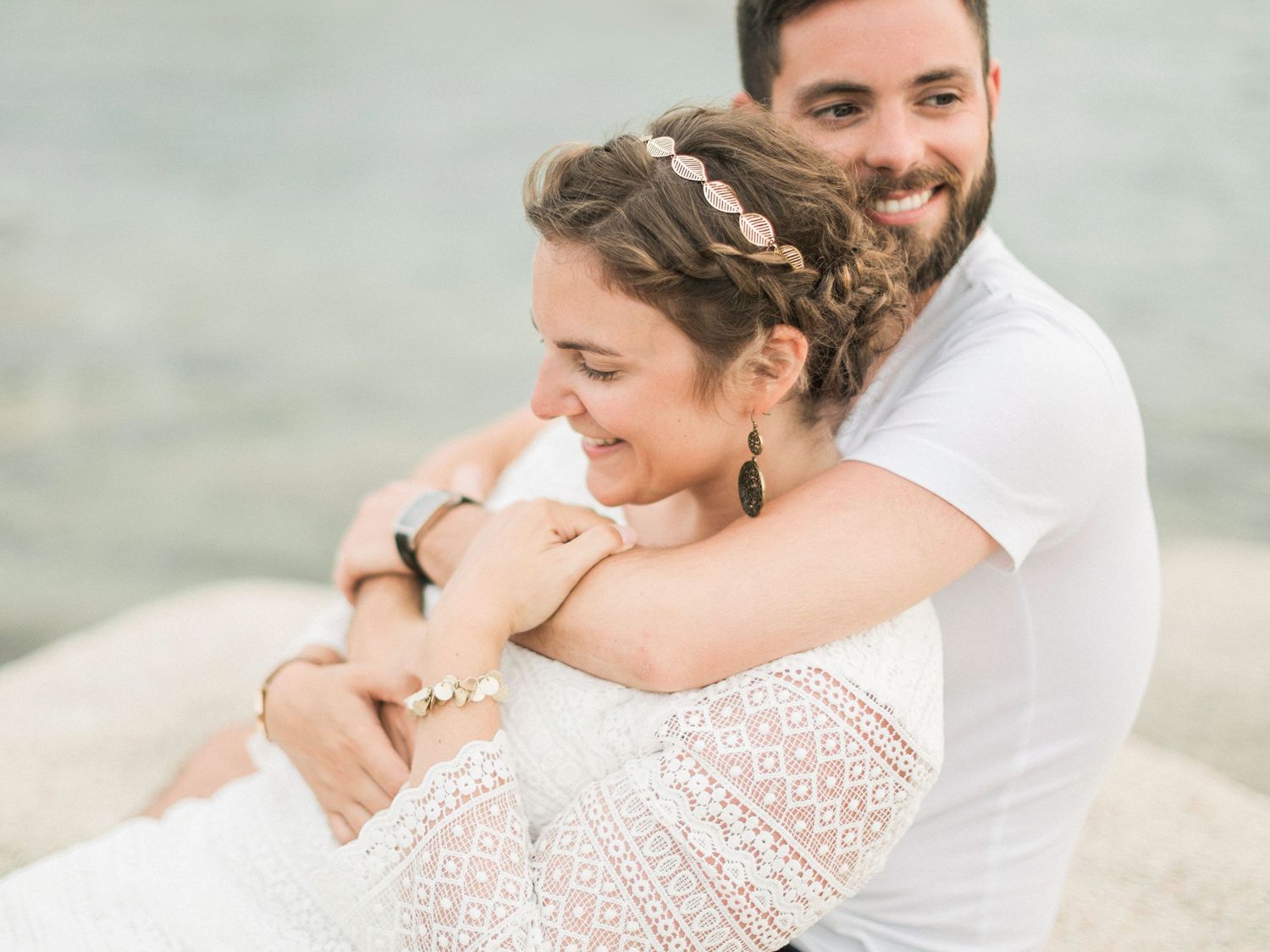 Un couple souriant s'enlaçant chaleureusement sur une plage rocheuse, la femme en robe de dentelle avec un bandeau fleuri.