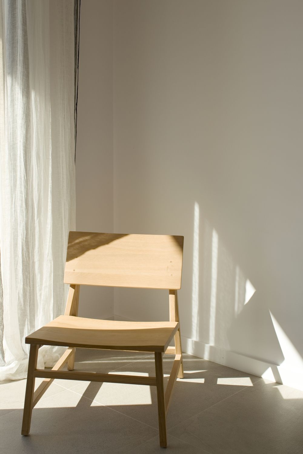 Une simple chaise en bois projette des ombres géométriques dans une pièce blanche baignée de soleil, aux rideaux de lin transparents.