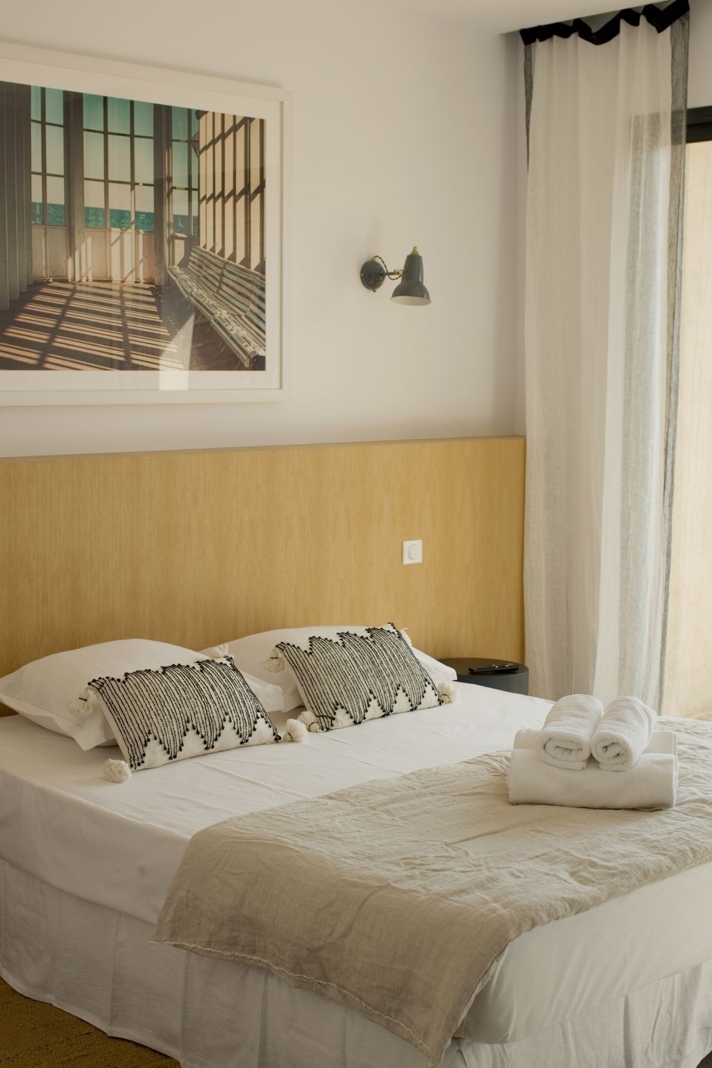 Chambre lumineuse et minimaliste avec tête de lit en bois, coussins à motifs, serviettes pliées et œuvres d'art architecturales encadrées.