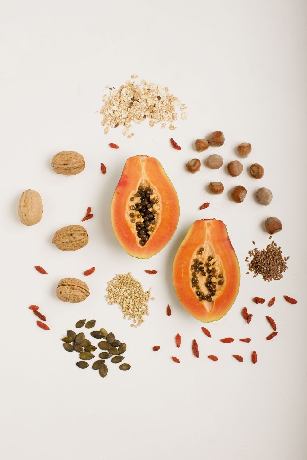 Papaye coupée en deux, entourée de noix, de graines, d'avoine et de baies de goji sur fond blanc en studio photo pour un livre de cuisine.