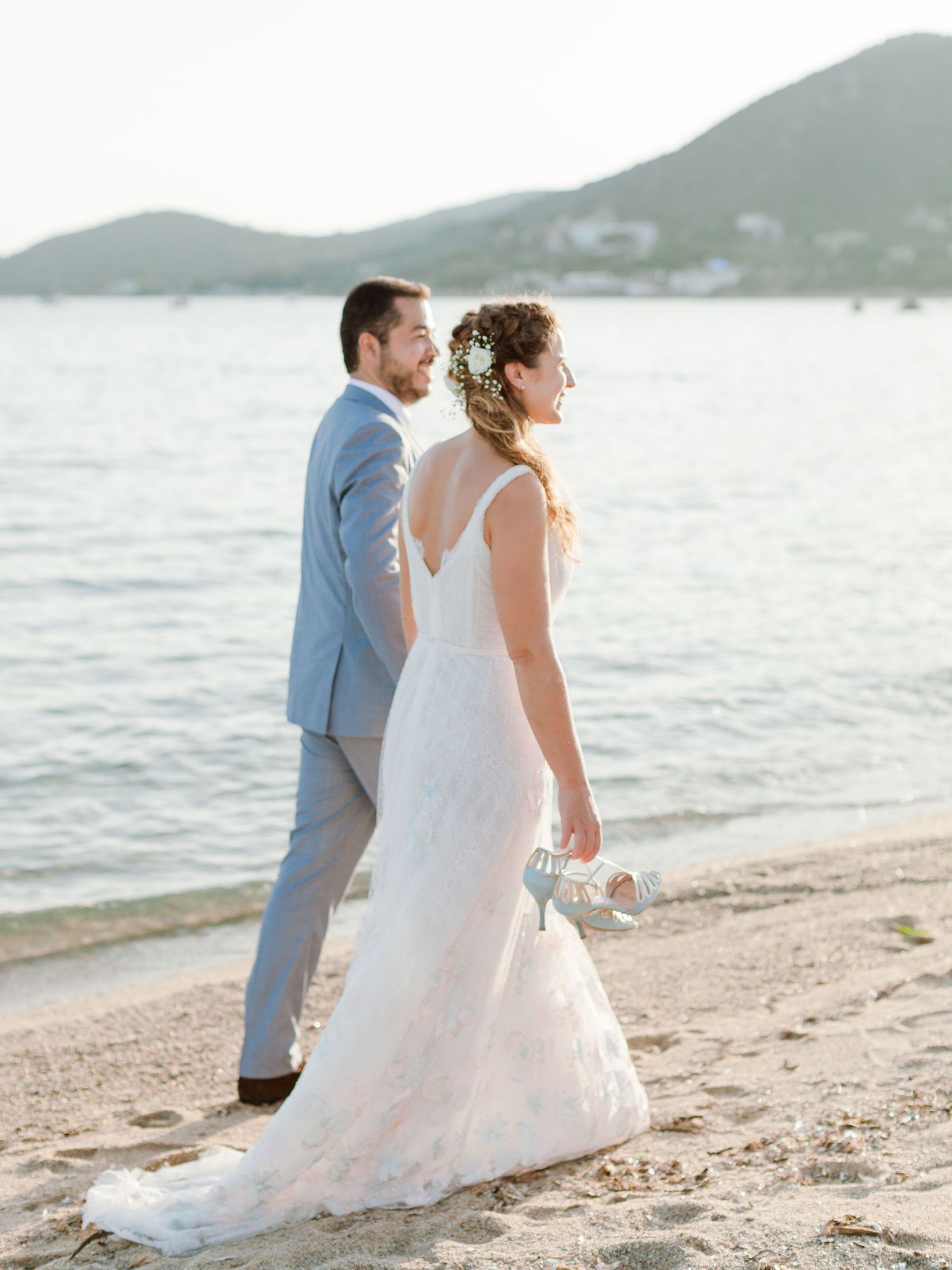 Les mariés marchent sur une plage de sable au coucher du soleil ; elle porte une robe en dentelle et lui un costume bleu clair