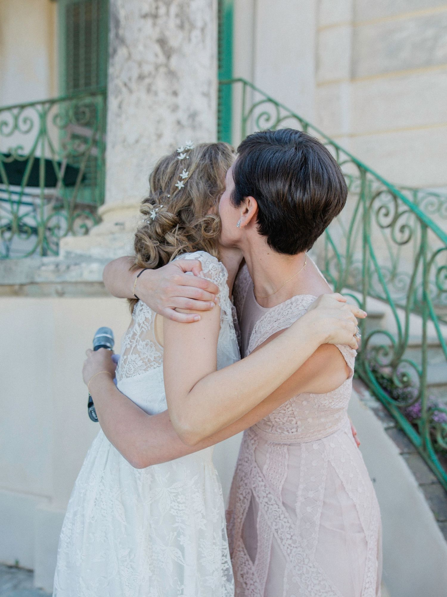 Two women in elegant lace dresses share an emotional embrace outside a vintage building with green iron railings.