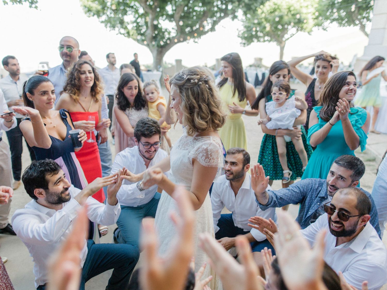 Joyful wedding guests dancing outdoors, surrounding the bride in a lively celebration circle at a sunny reception.