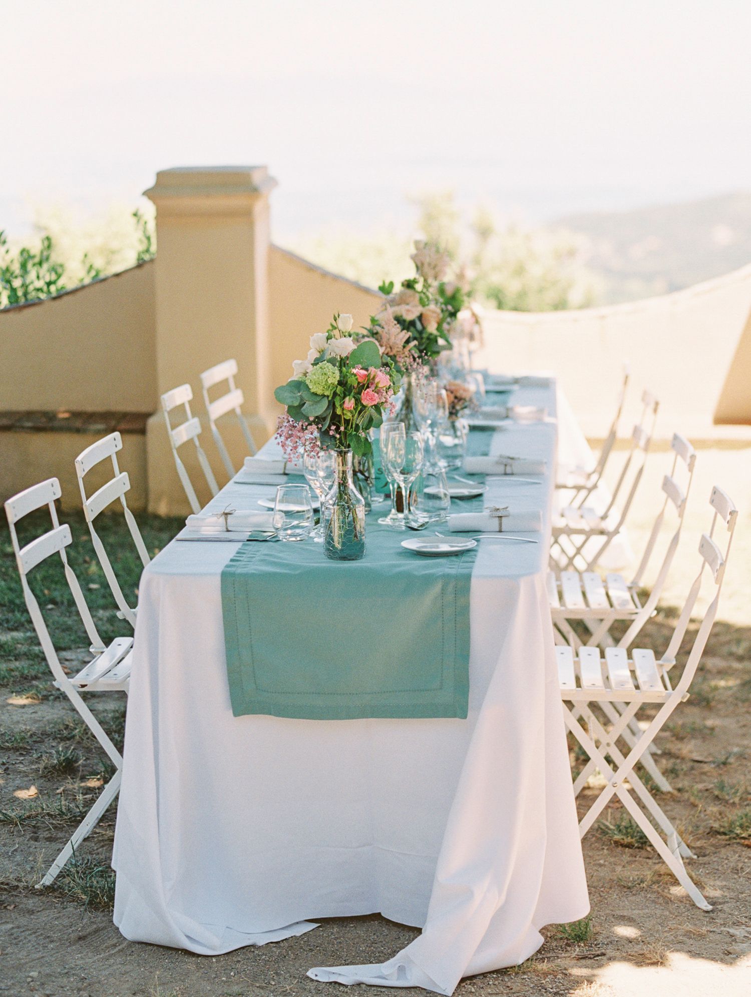 Elegant outdoor wedding reception table with teal runner, white chairs, floral centerpieces, and scenic hillside backdrop.