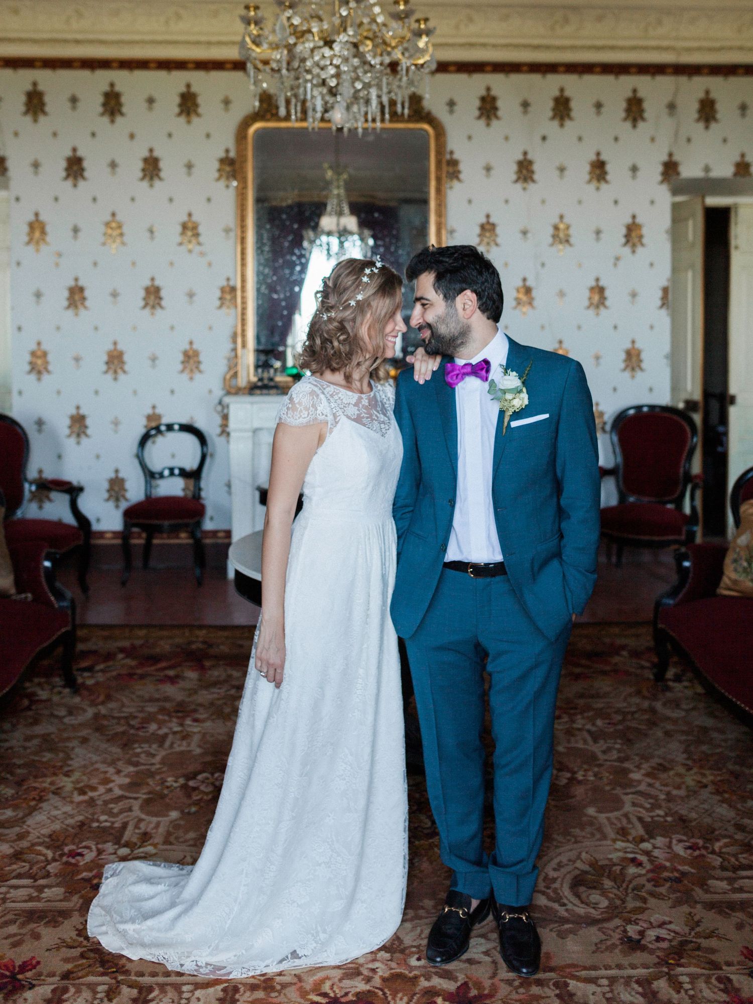 Bride in lace gown and groom in teal suit with purple bow tie share a romantic moment in an elegant ballroom.