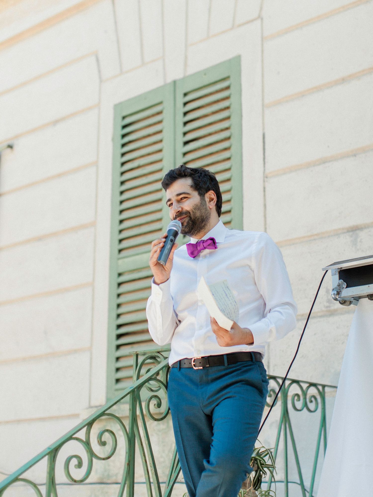 Man in white shirt and purple bow tie speaks into microphone at elegant outdoor venue with green shutters.