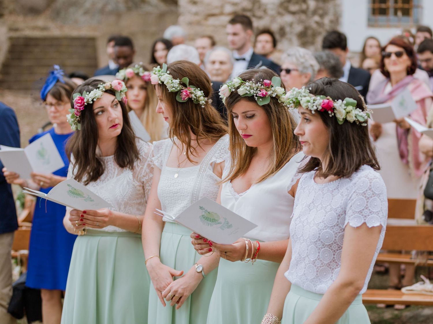 Des demoiselles d'honneur en jupes menthe et couronnes de fleurs lisent des documents lors d'une cérémonie de mariage en plein air.