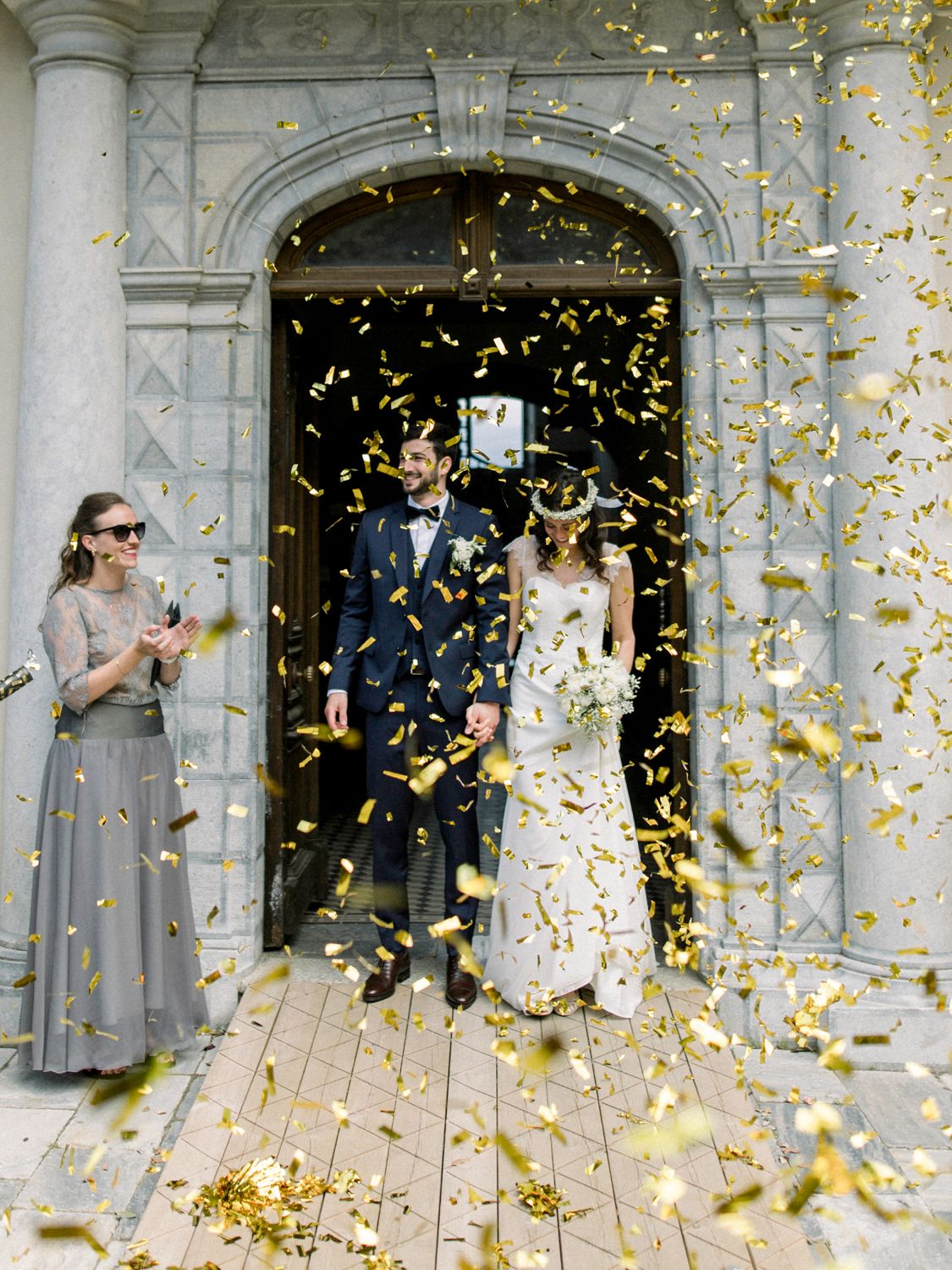 Newlywed couple exits church amid golden confetti shower, with bridesmaid in gray gown celebrating beside them.