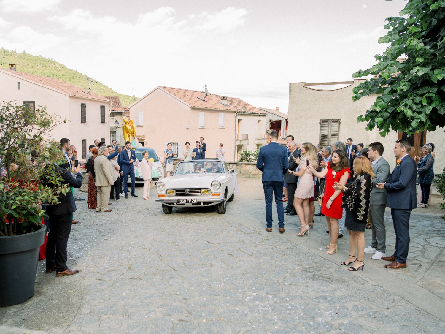 Guests gather in a sunlit village square as a vintage convertible car arrives at an outdoor wedding celebration.