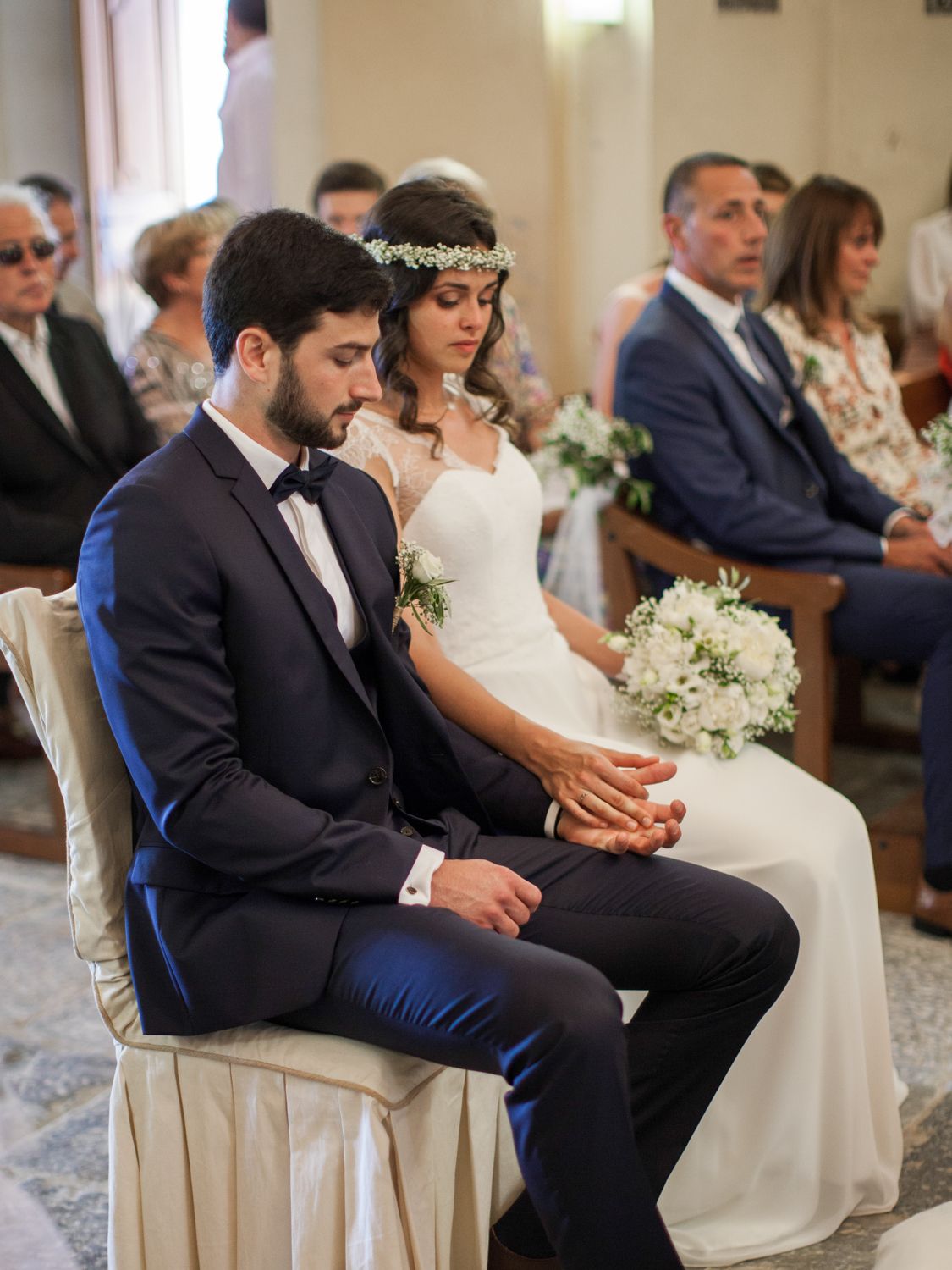 Bride and groom sit together during wedding ceremony, bride wearing floral crown holding white bouquet.