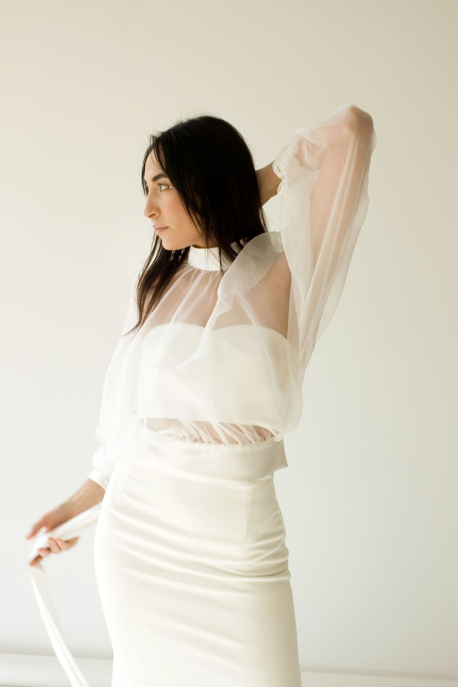 Une mariée élégante, vêtue d'un haut en organza blanc by romance  et d'une jupe en satin, pose avec grâce sur un fond clair.