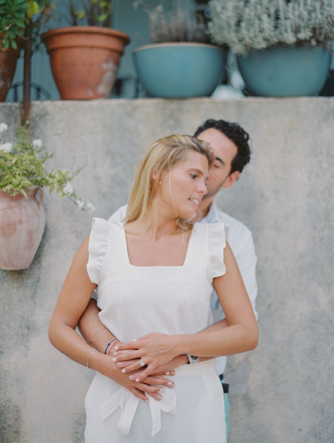 Un couple s'enlace tendrement à l'extérieur, la femme en robe blanche à volants, l'homme en chemise claire, avec des pots en terre cuite