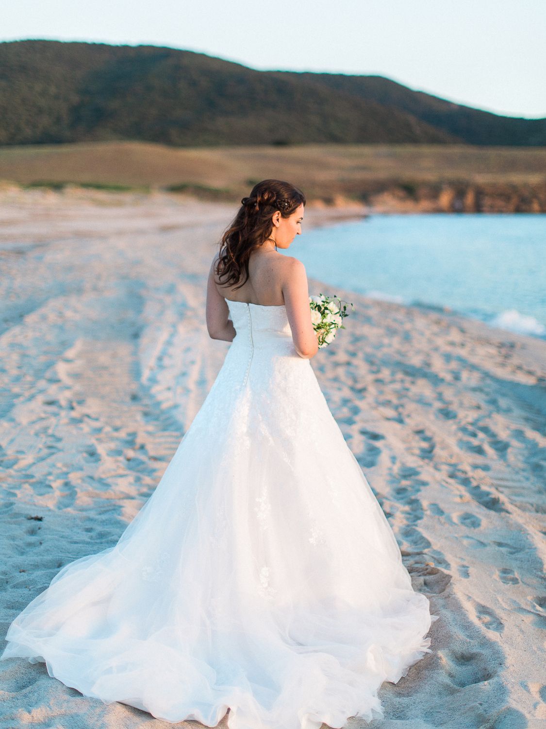 Mariée en robe de bal blanche sans bretelles tenant un bouquet sur une plage de sable au coucher du soleil