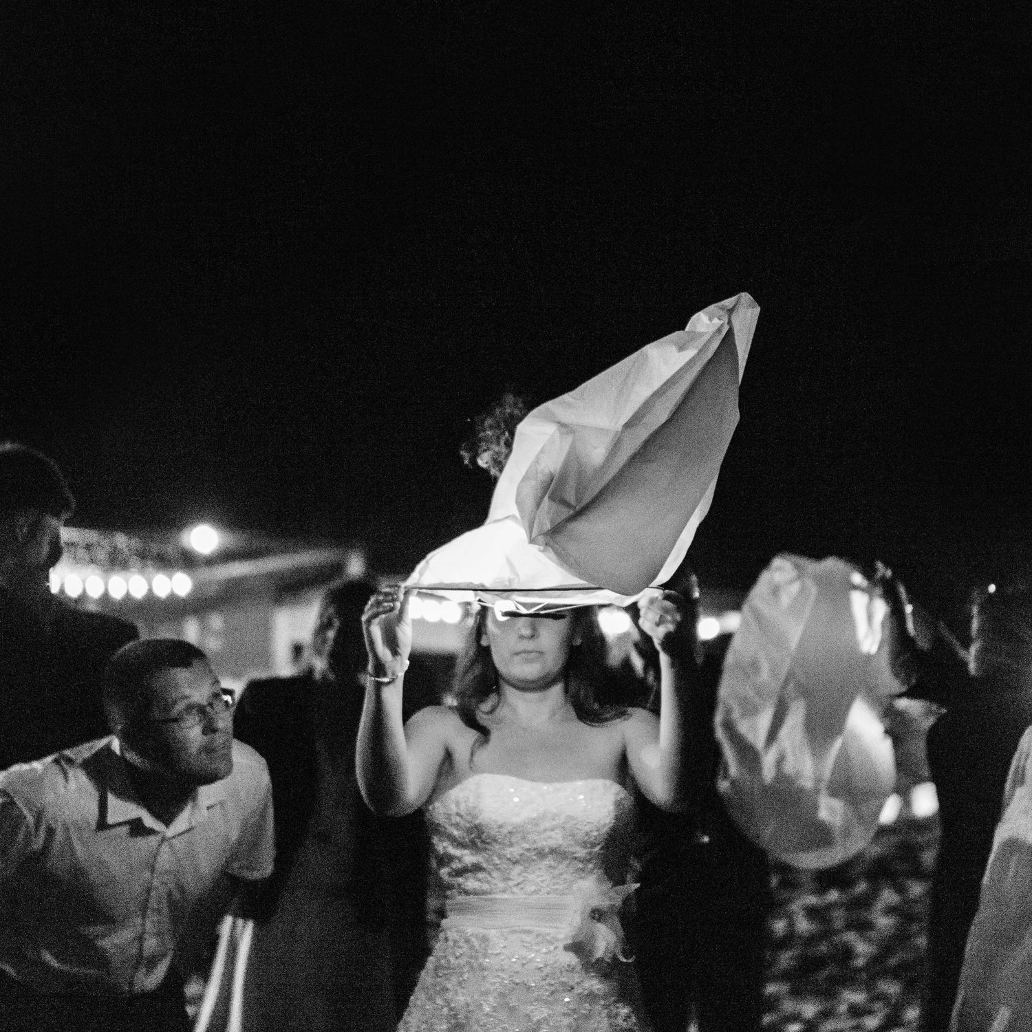 Une mariée en robe de mariée lâche une lanterne céleste lors d'une célébration nocturne en plein air, photographiée en noir et blanc.