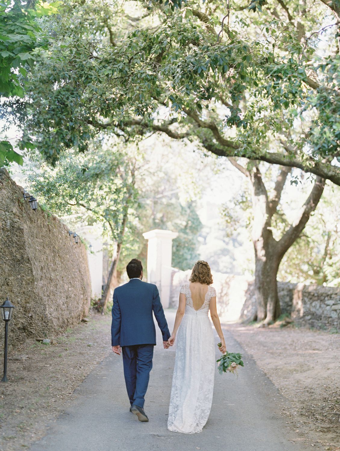 Les mariés marchent main dans la main sur une allée bordée d'arbres, la mariée tenant un bouquet vert. Au Palazzu Nicrosi cap Corse.