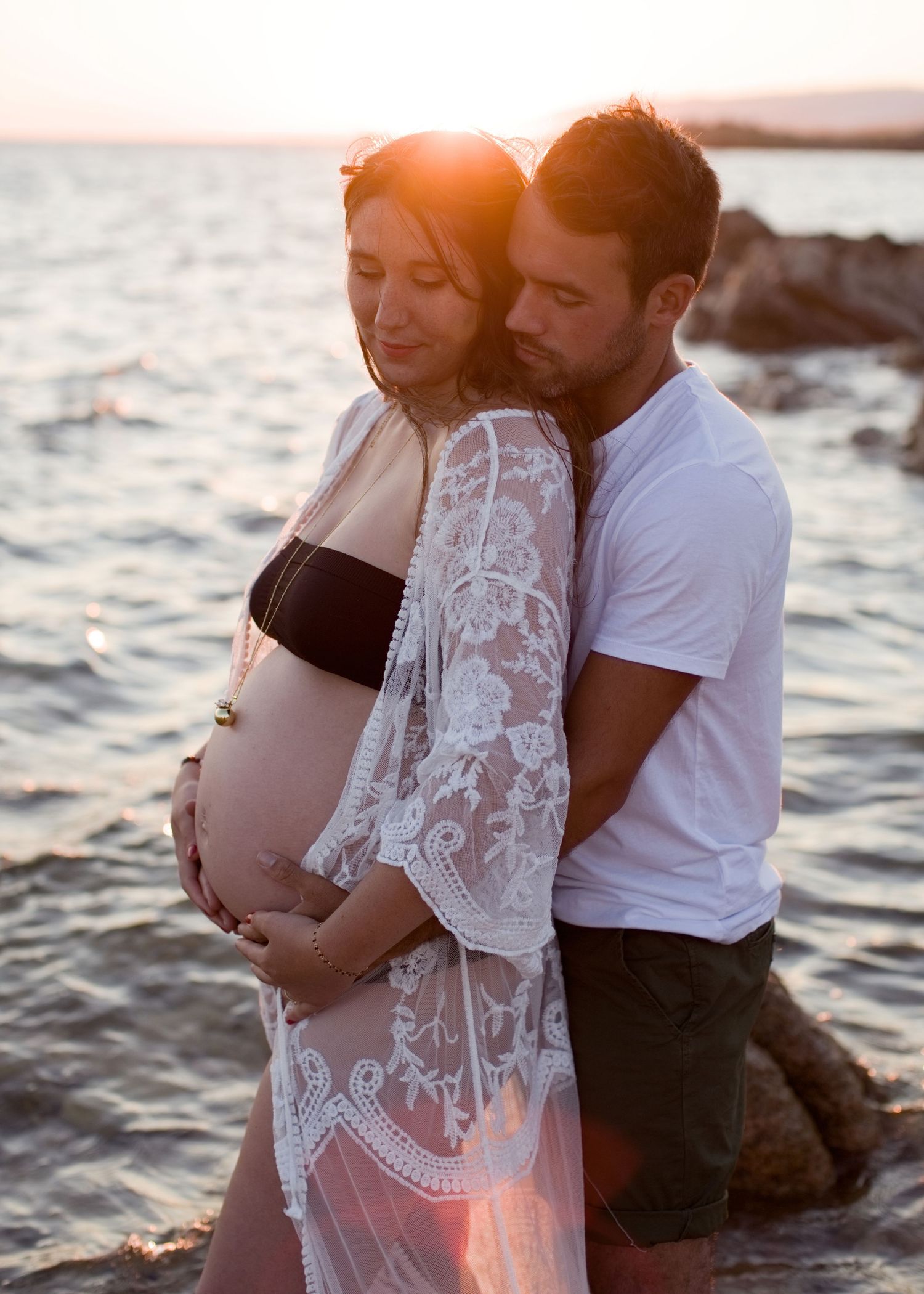 Un couple partage un tendre moment de maternité sur une plage rocheuse au coucher du soleil, sous un soleil doré et chaleureux à Bonifacio.