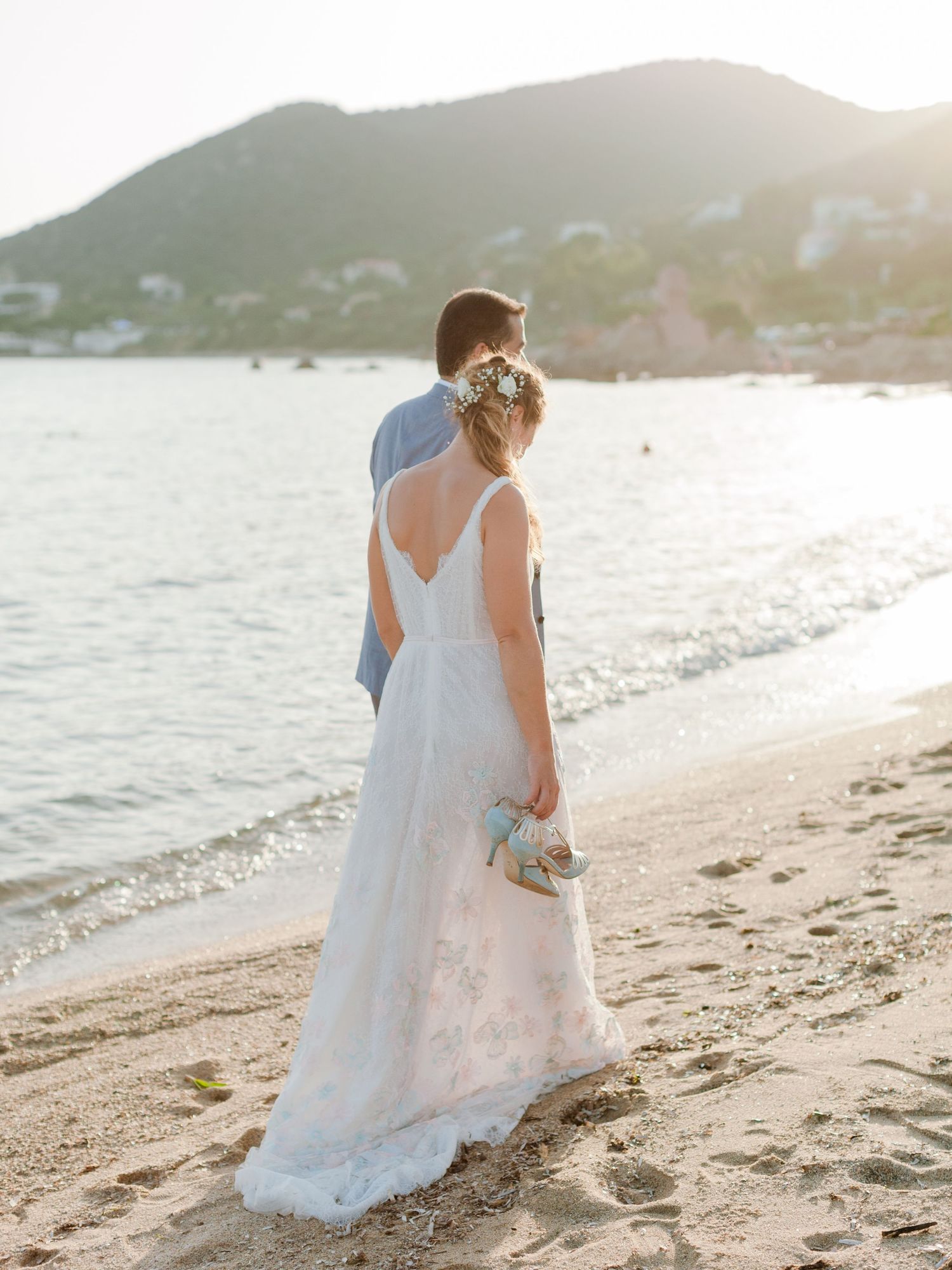 Une mariée en robe blanche et un marié se tiennent sur une plage de sable au coucher du soleil, avec la montagne et l'océan en toile de fond