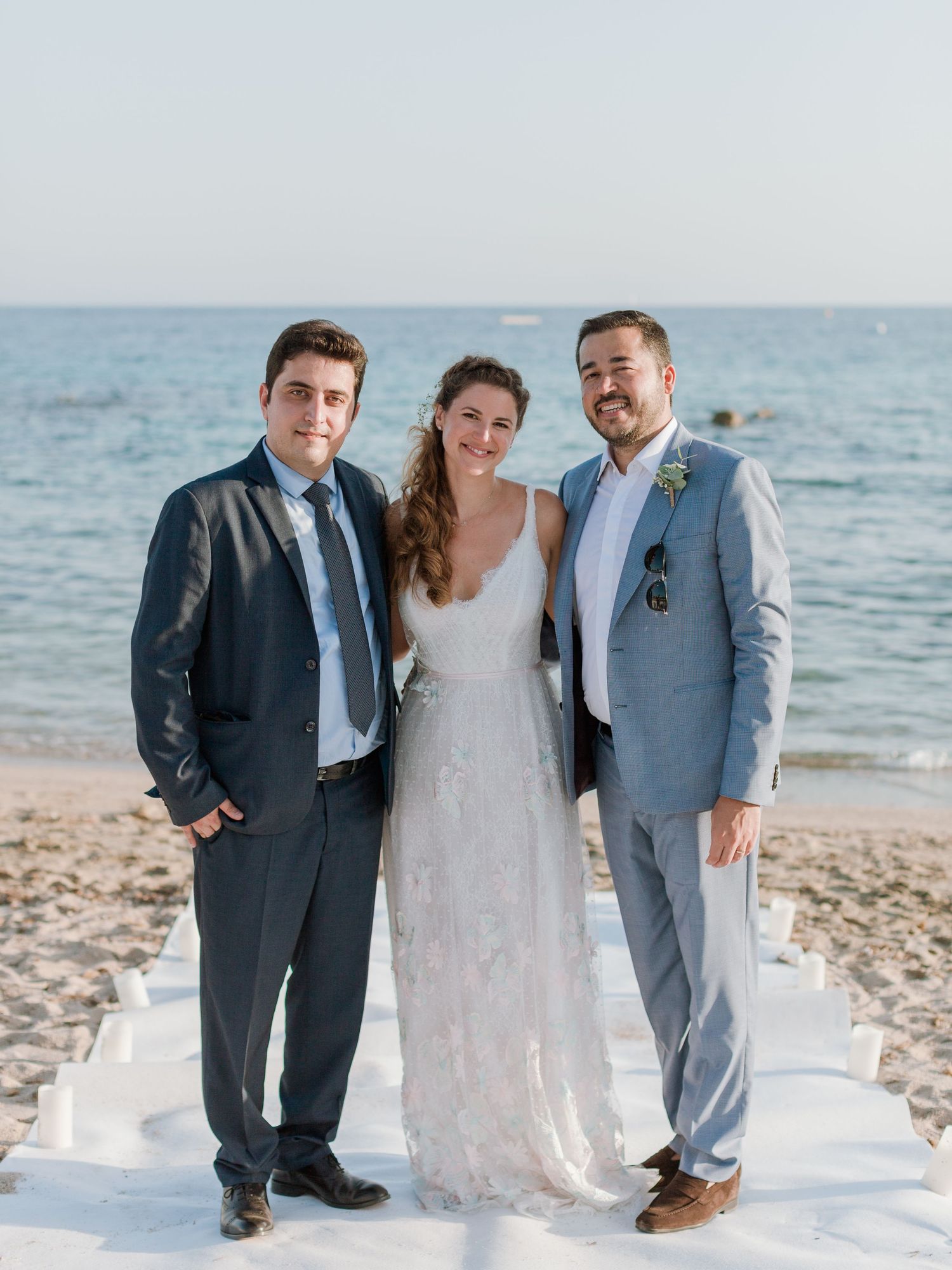 Une mariée en robe de dentelle blanche pose avec deux hommes en costume, dans le cadre d'une cérémonie de mariage sur une plage ensoleillée.