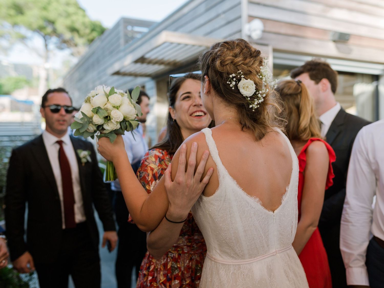 Une mariée en robe blanche dos nu reçoit des félicitations tout en tenant un bouquet lors d'une réception de mariage en plein air.