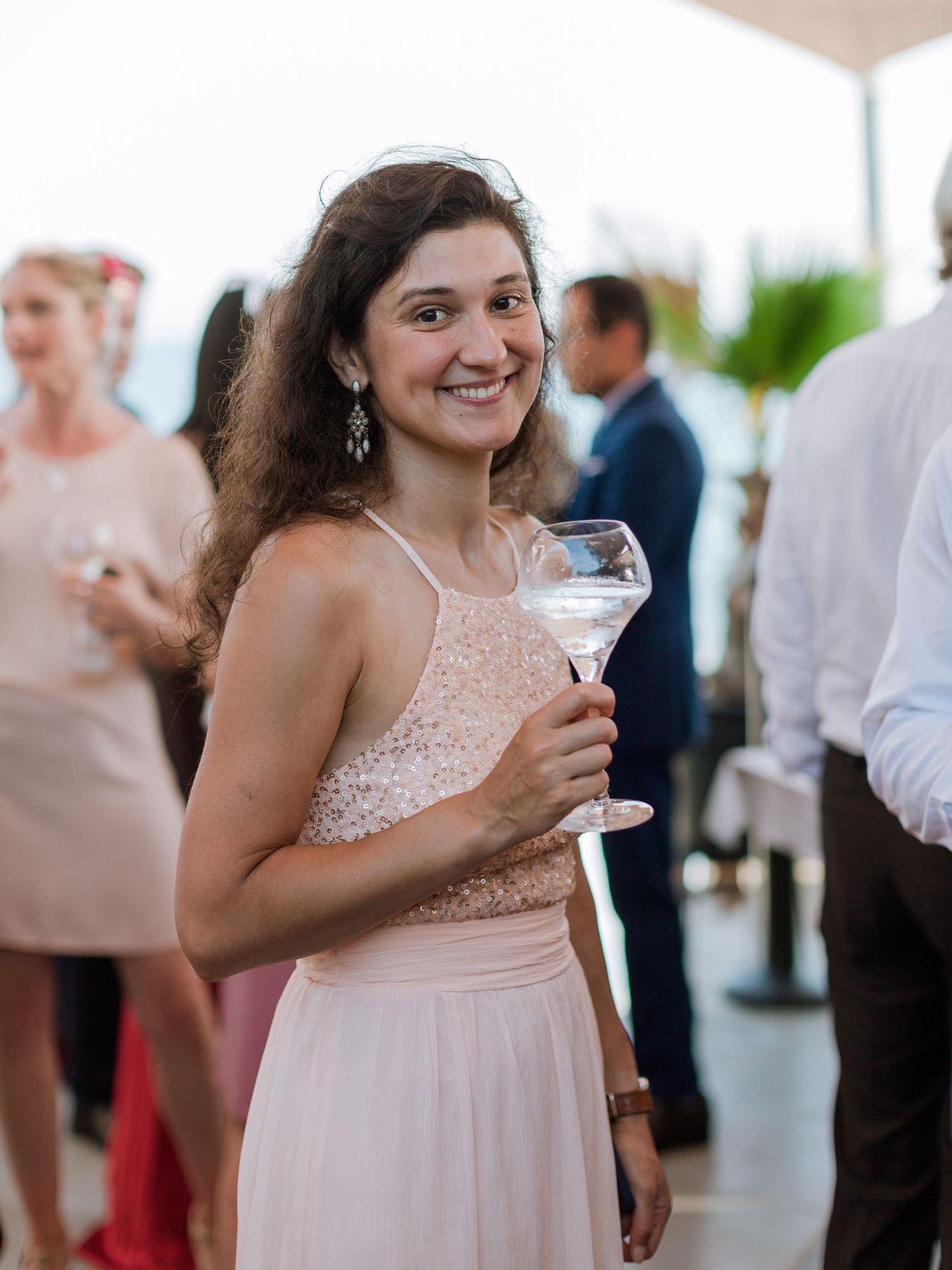 Jeune femme en robe rose souriant lors d'une fête, tenant un verre de martini à un événement mondain élégant en plein air.