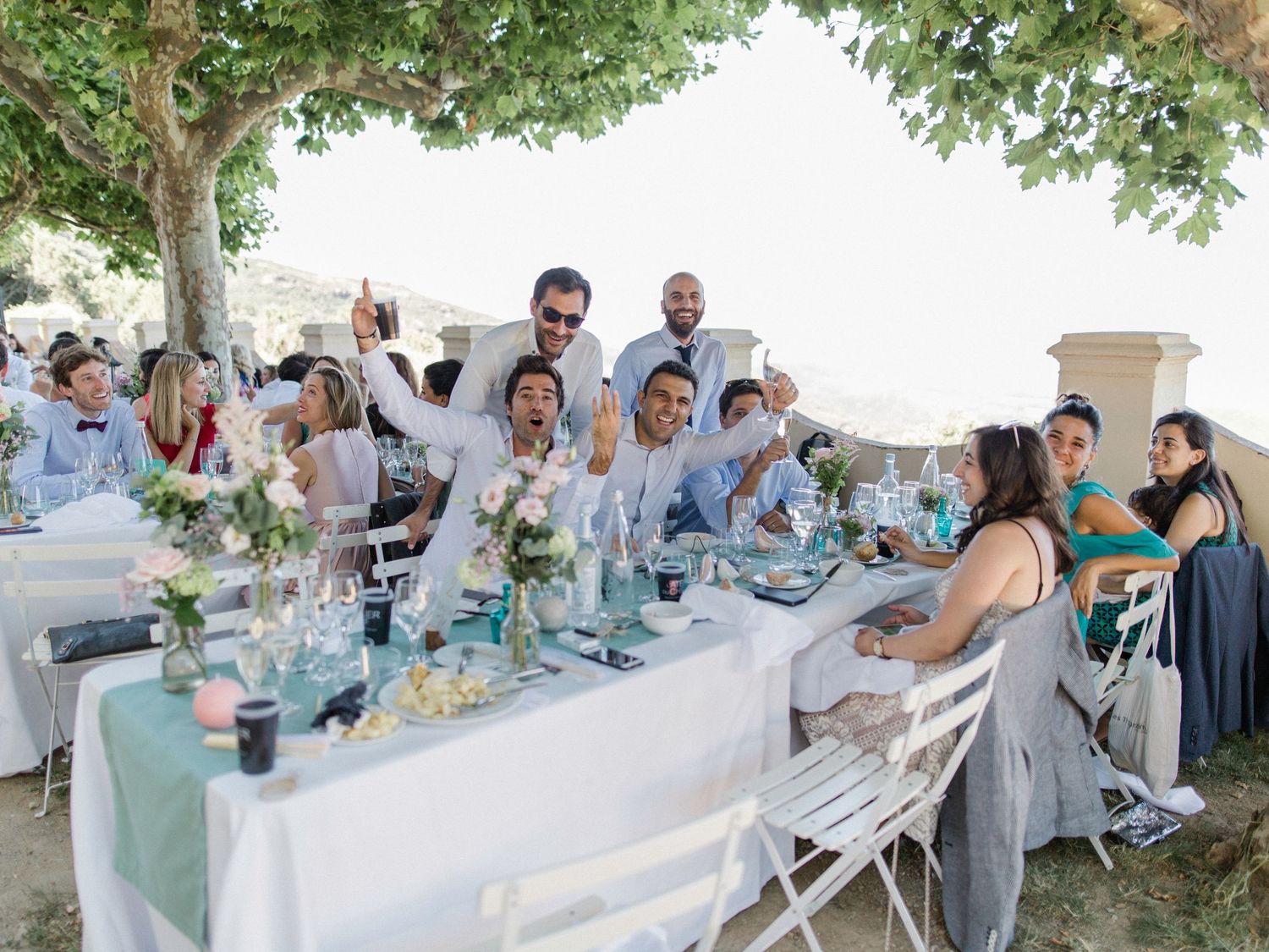 Guests celebrate at an outdoor wedding reception, toasting at a white-draped table under a tree.