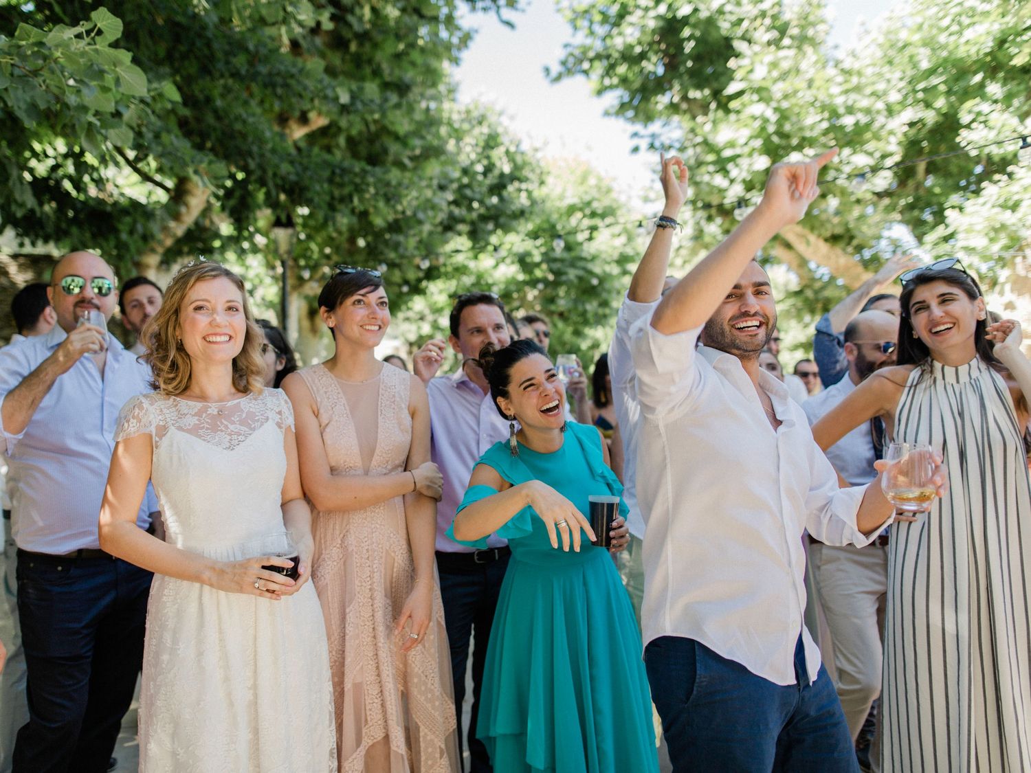 Joyful wedding guests celebrating outdoors, bride in white lace dress, friends cheering and raising hands in sunny garden.