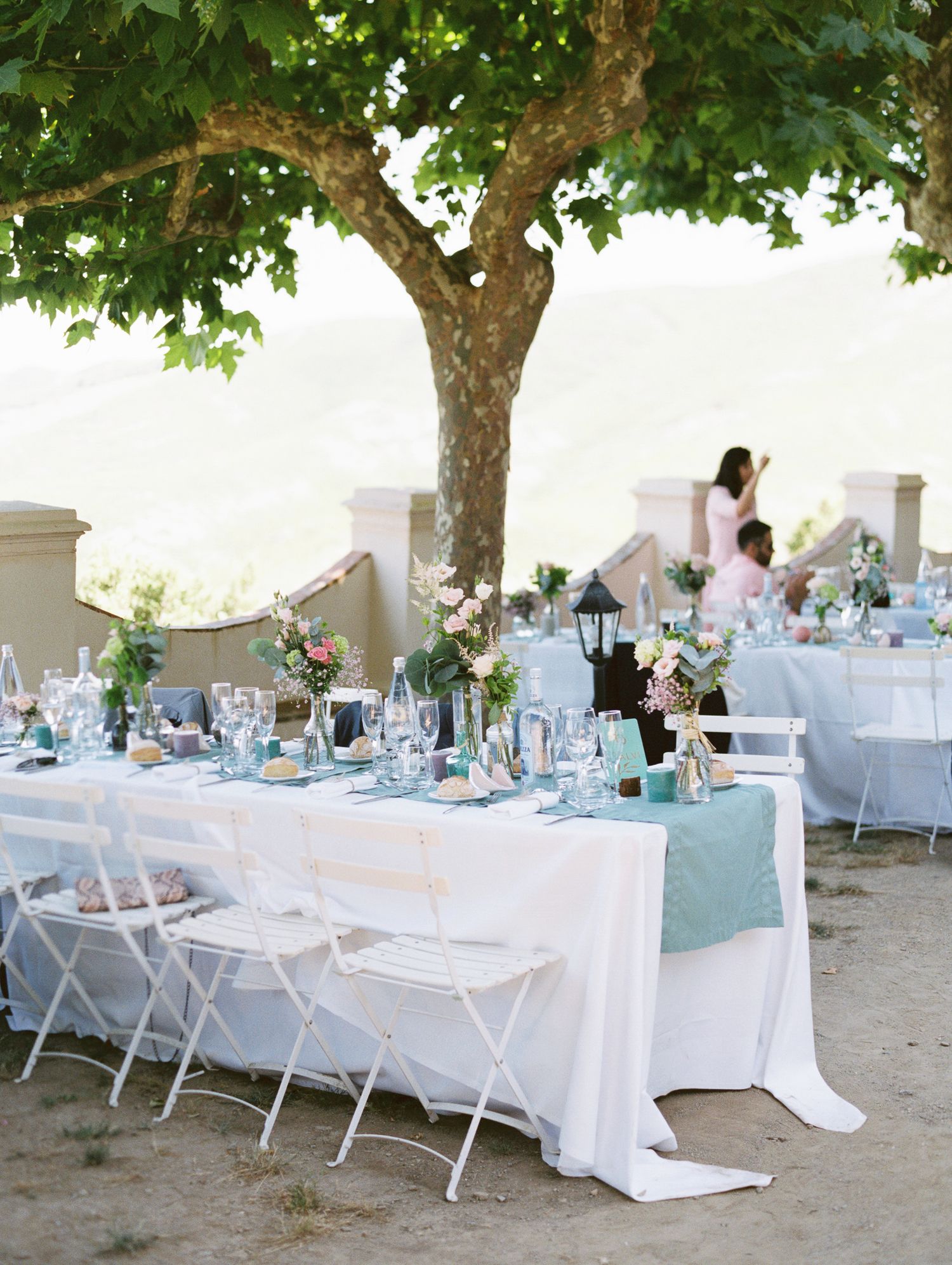 Elegant outdoor wedding reception with white tablecloths, teal runners, floral centerpieces, and white chairs under a tree.