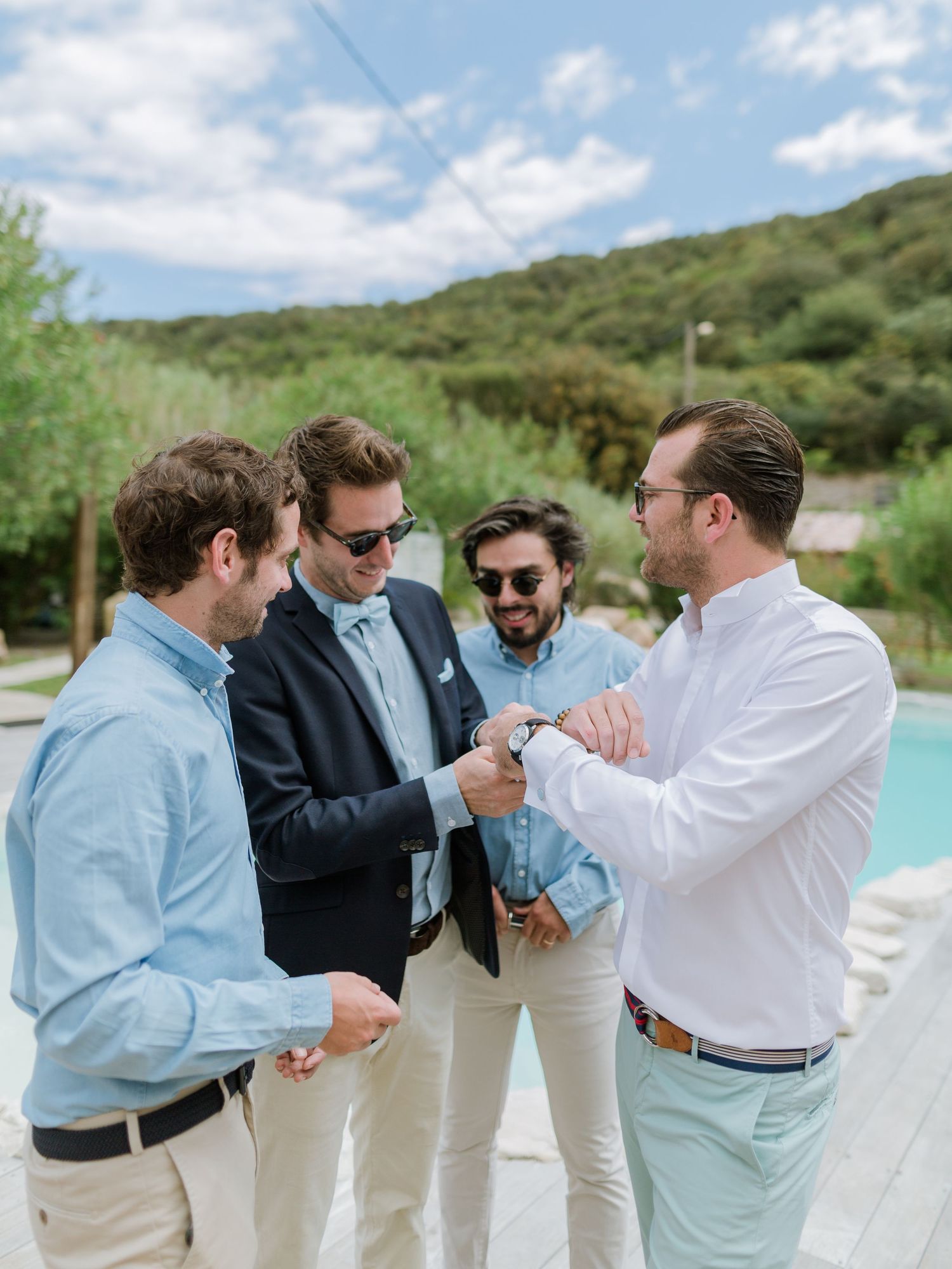 Quatre hommes élégants réunis au bord d'une piscine, l'un d'eux ouvrant une bouteille, vêtus d'une tenue décontractée chic en extérieur.
