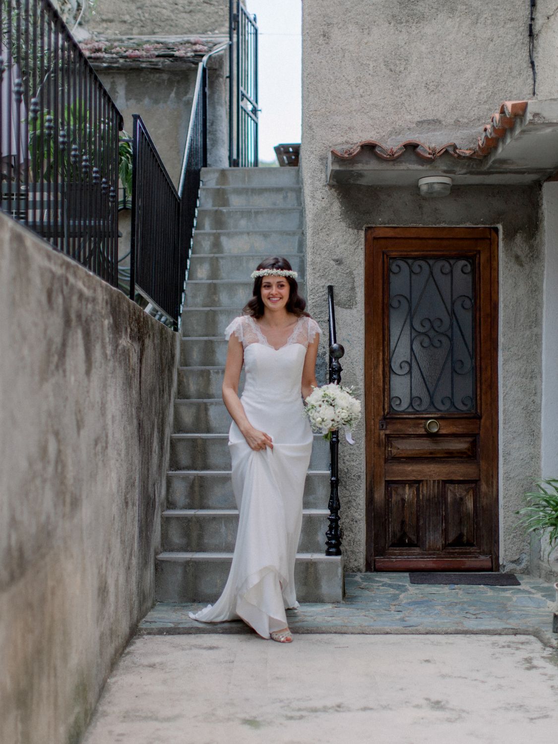 Smiling bride in white gown holding bouquet stands by ornate wooden door with wrought iron staircase backdrop.